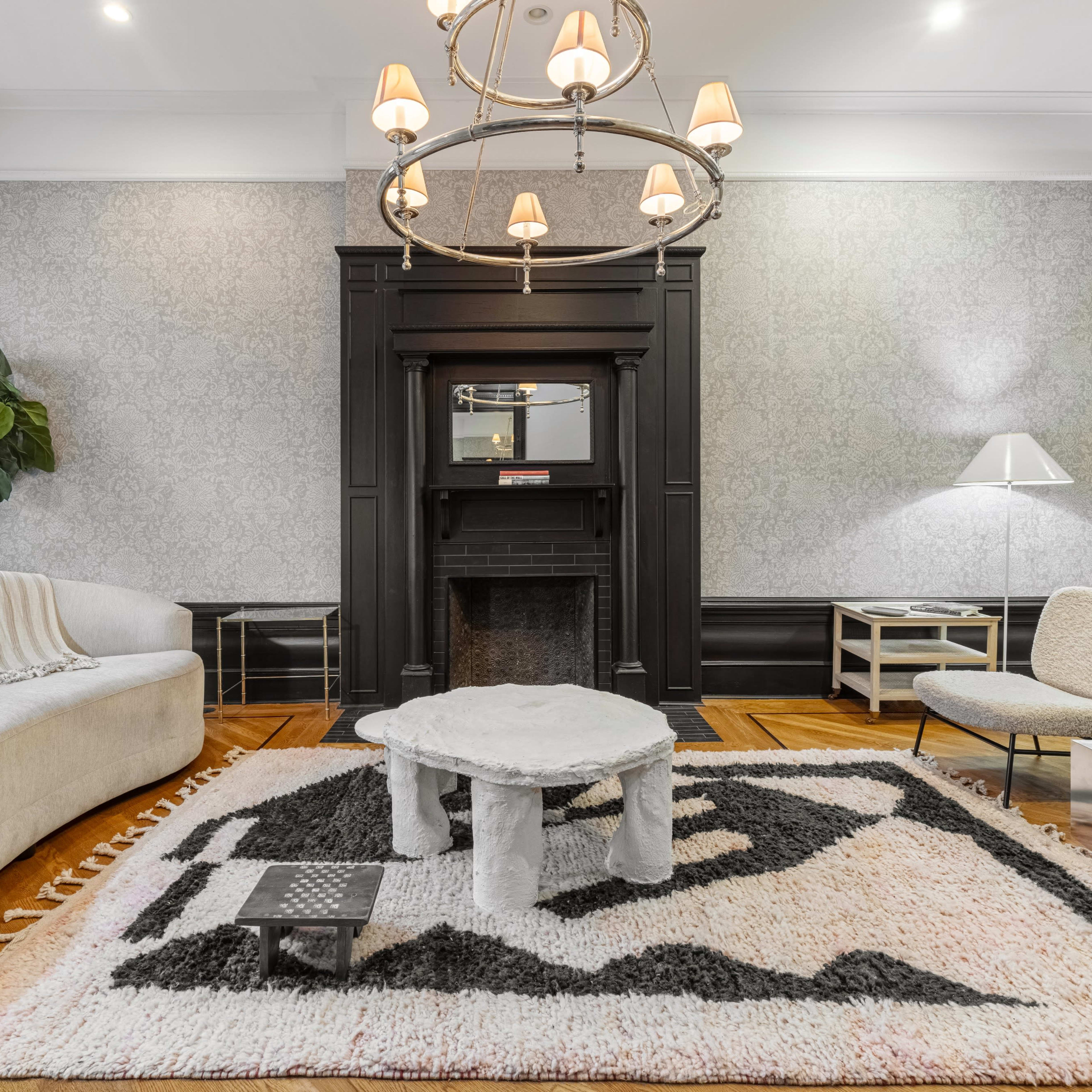 A living room featuring a plush gray sofa, a large patterned area rug, a marble coffee table, and a striking chandelier above an elegant fireplace.