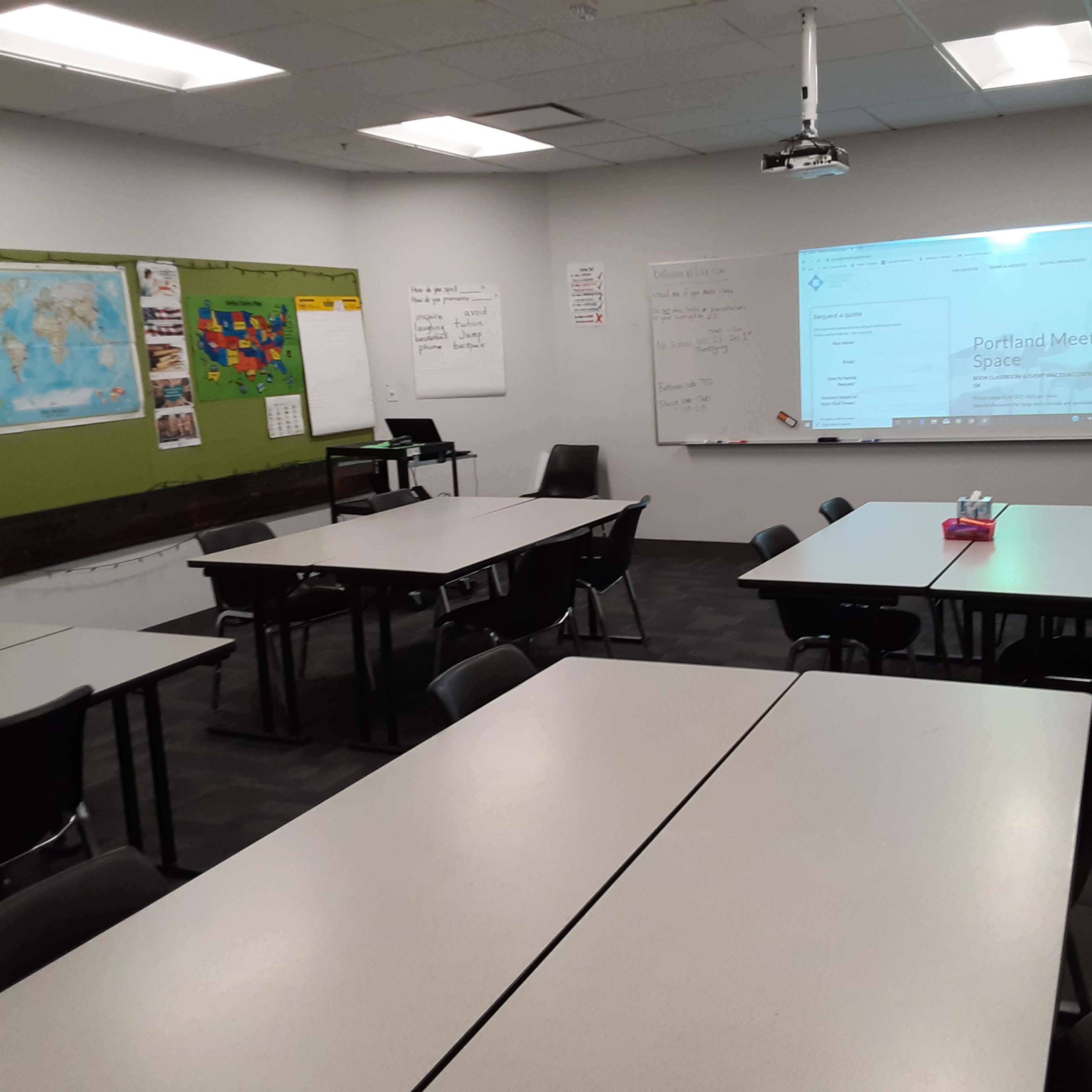 A classroom is arranged with several tables and chairs, featuring a projector and whiteboard on one wall along with a map.