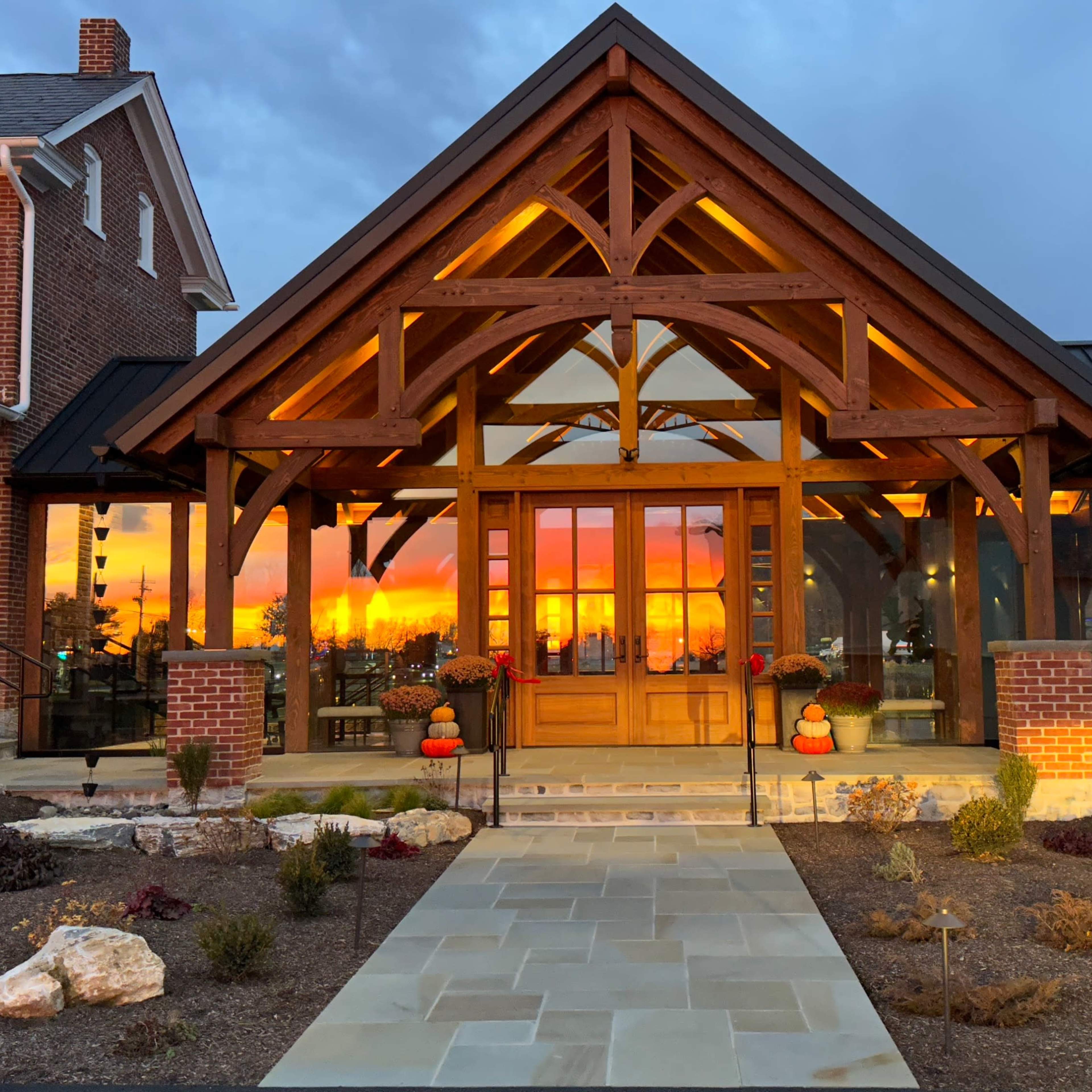A large wooden-framed entrance of a house with glass panels, flanked by two buildings, as a sunset casts warm colors in the sky.