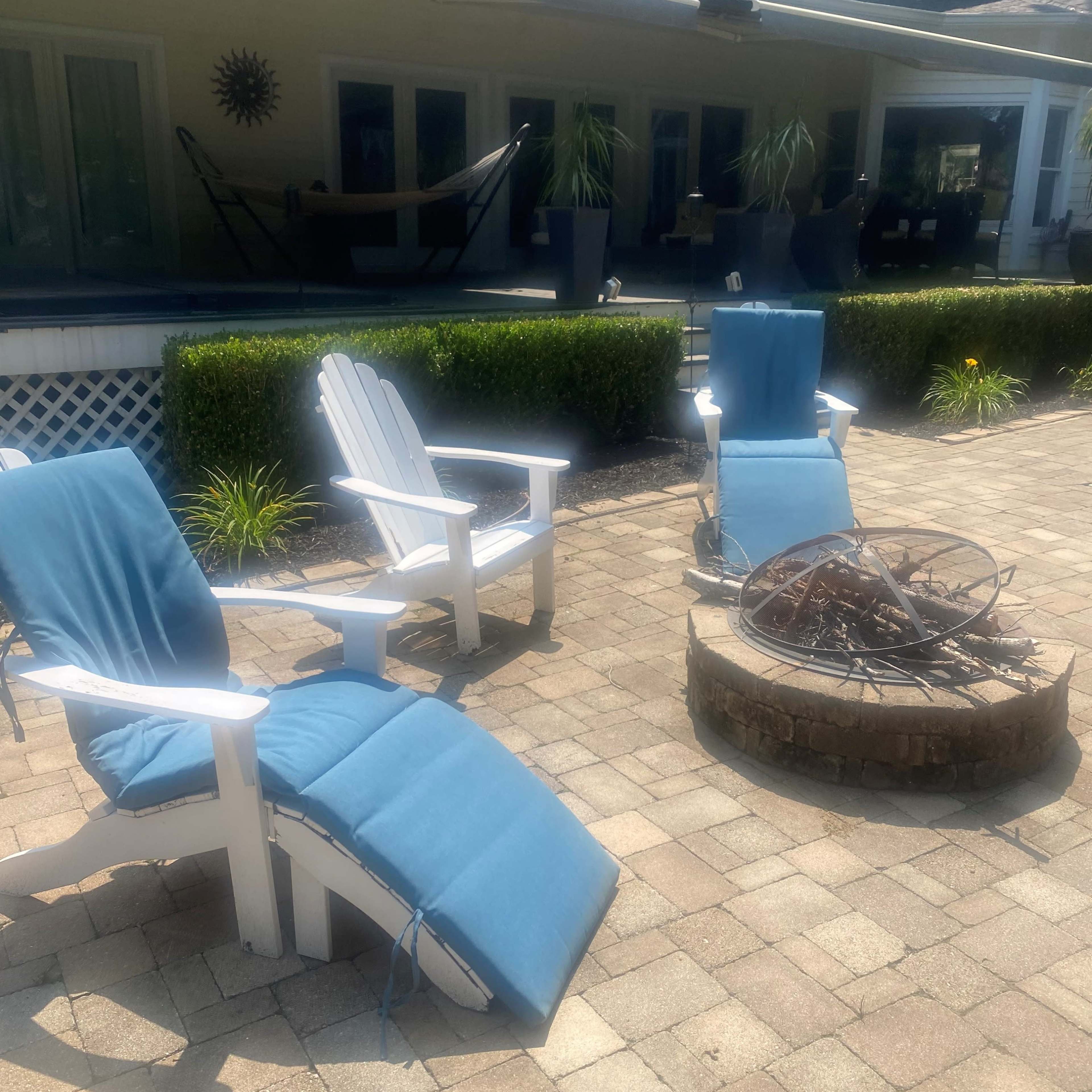 Two white lounge chairs with blue cushions positioned near a circular fire pit on a stone patio.