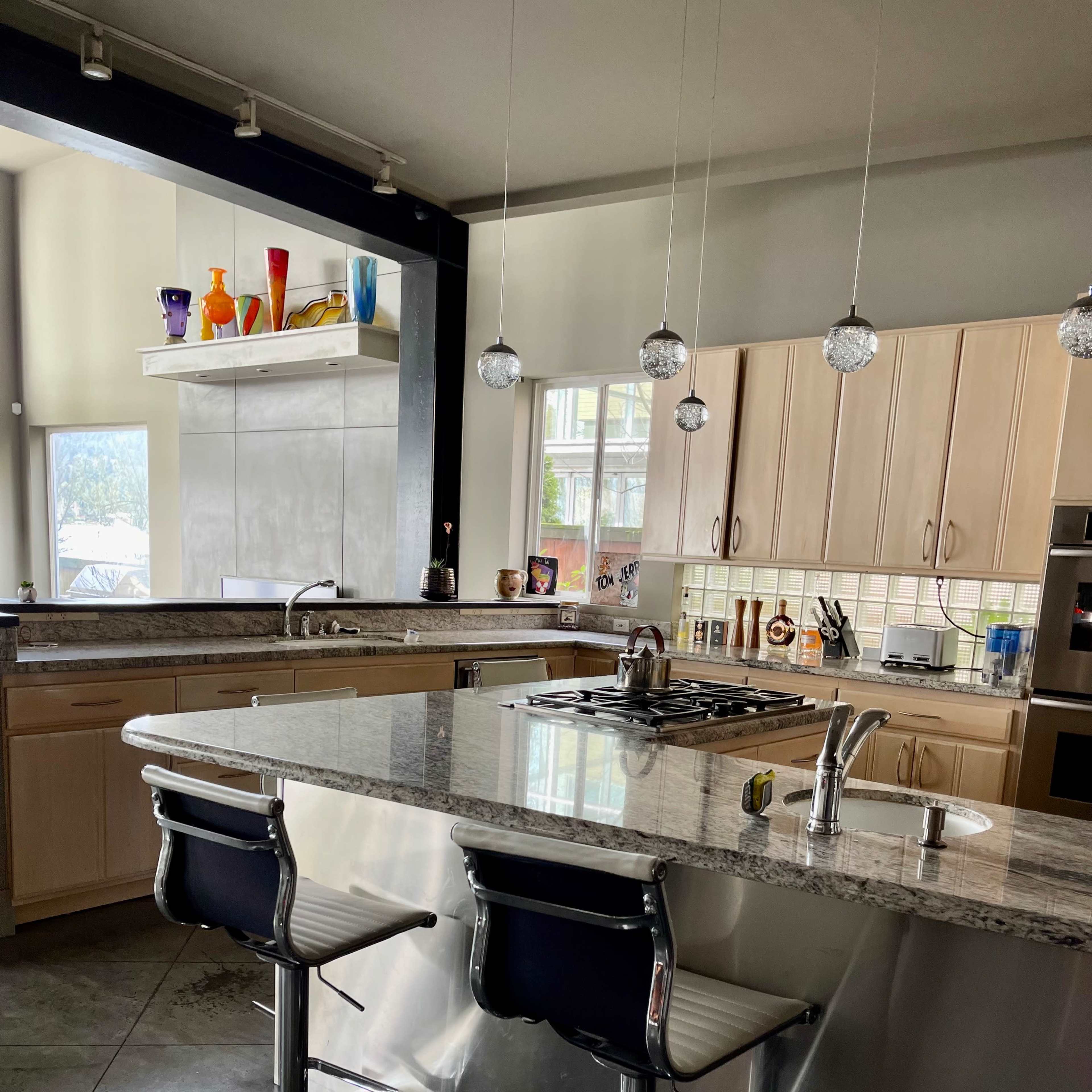 A modern kitchen featuring a large island with two barstools, stainless steel appliances, and a range of colorful decorations on the counter.