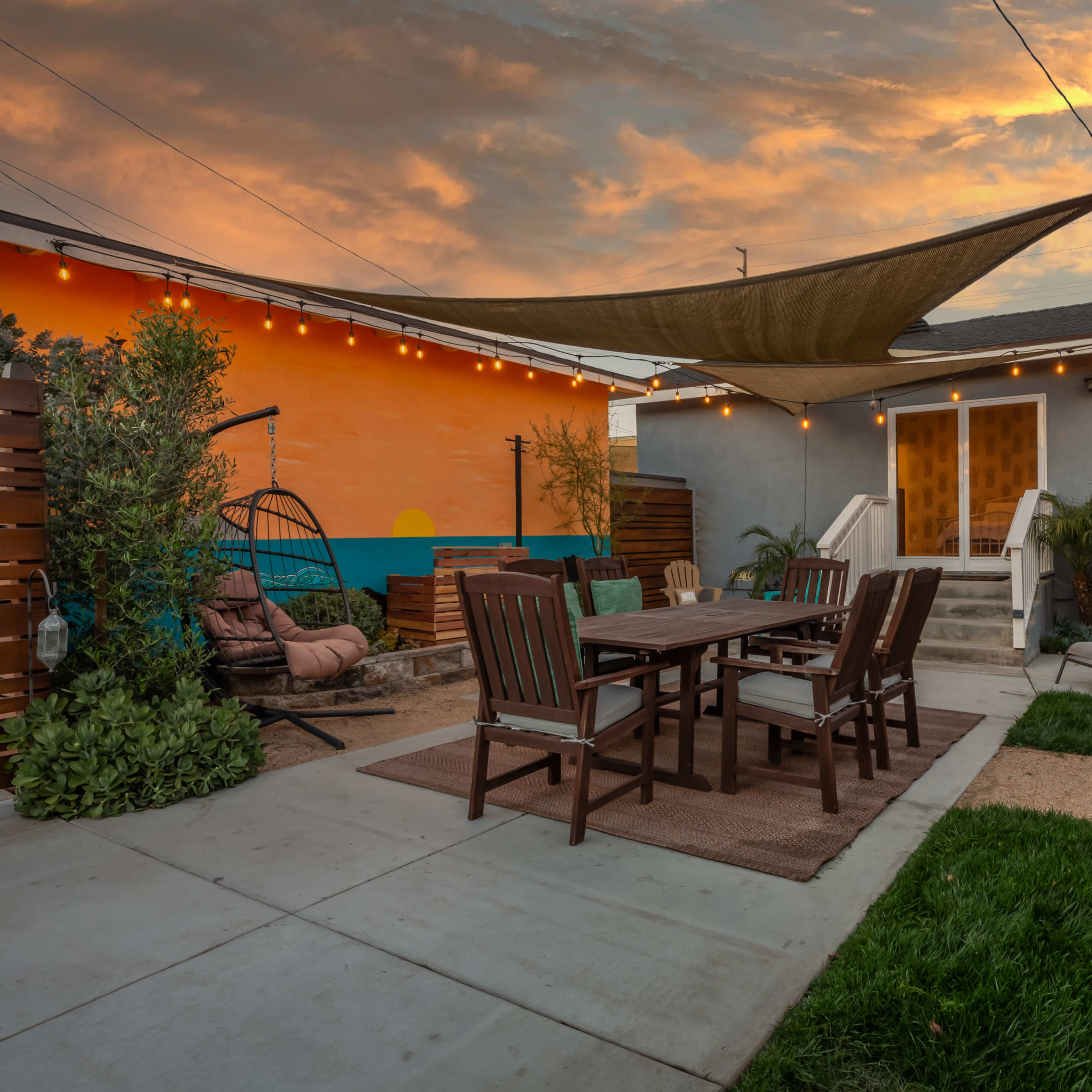 A patio with a wooden dining set, lounge chairs, and string lights, set against a colorful mural and a sunset sky.