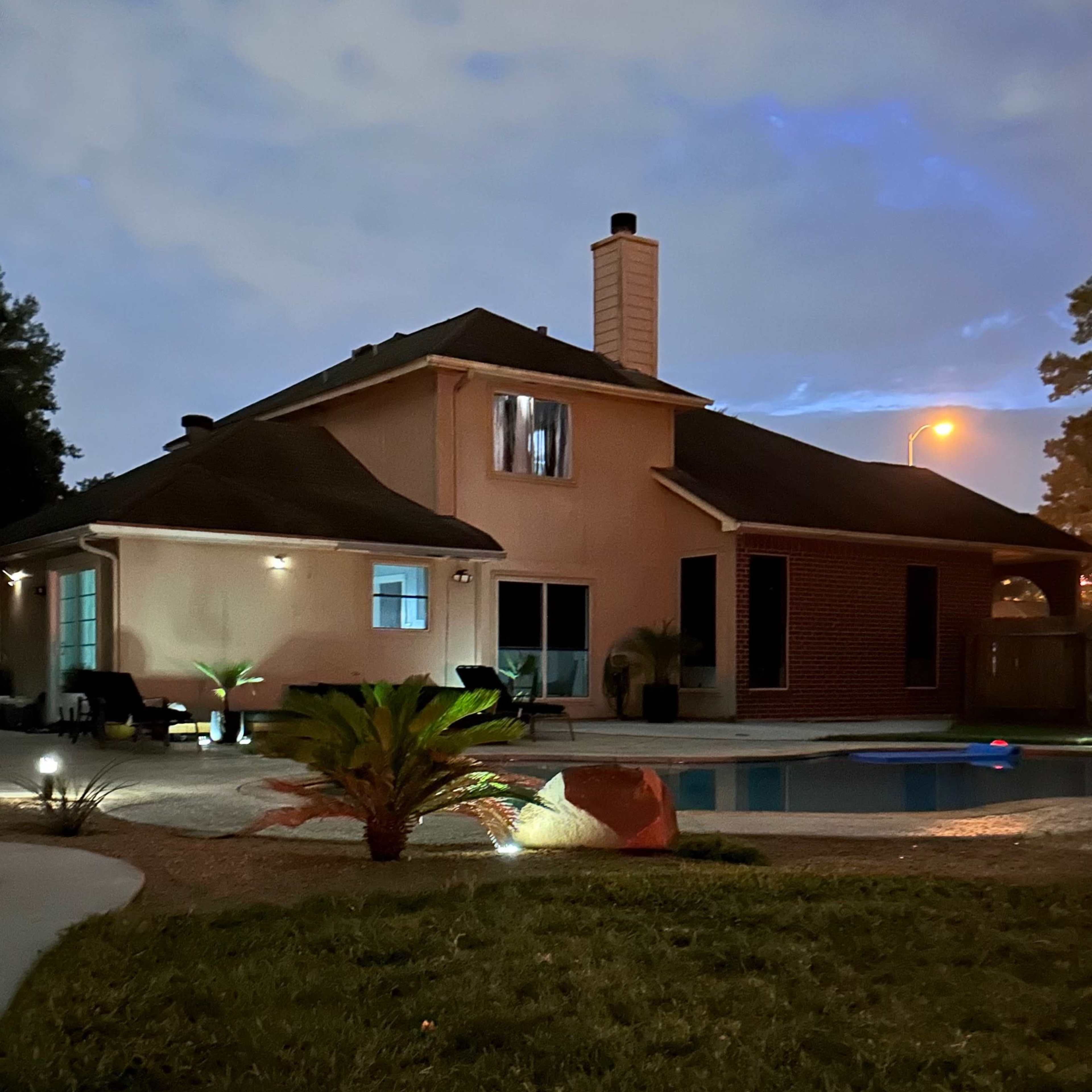 A two-story house with a swimming pool is illuminated in the evening, featuring a mix of landscaping and outdoor lighting.