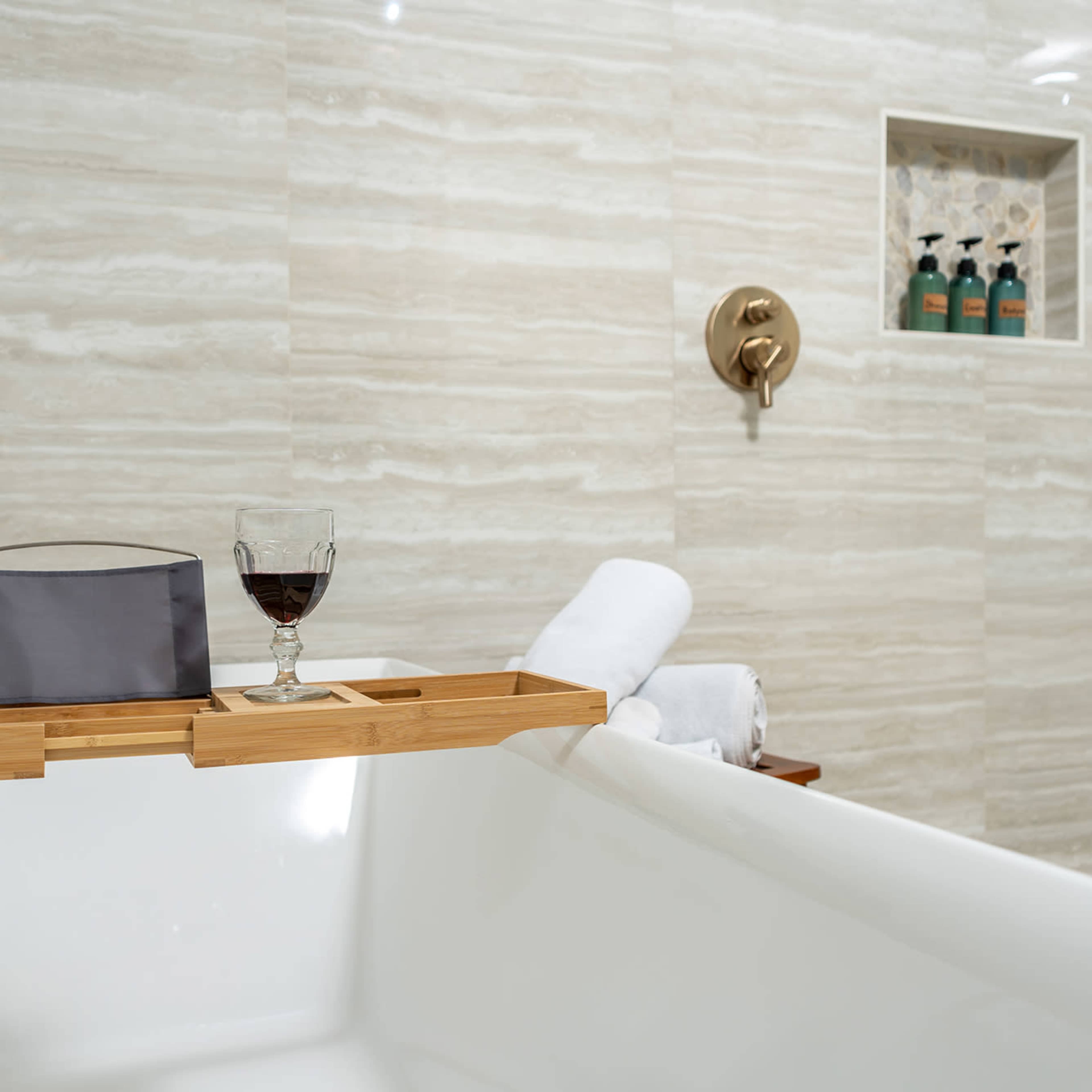 A modern bathroom features a white bathtub with a wooden tray holding a glass of dark liquid and towels, set against striped marble walls and a niche displaying toiletries.