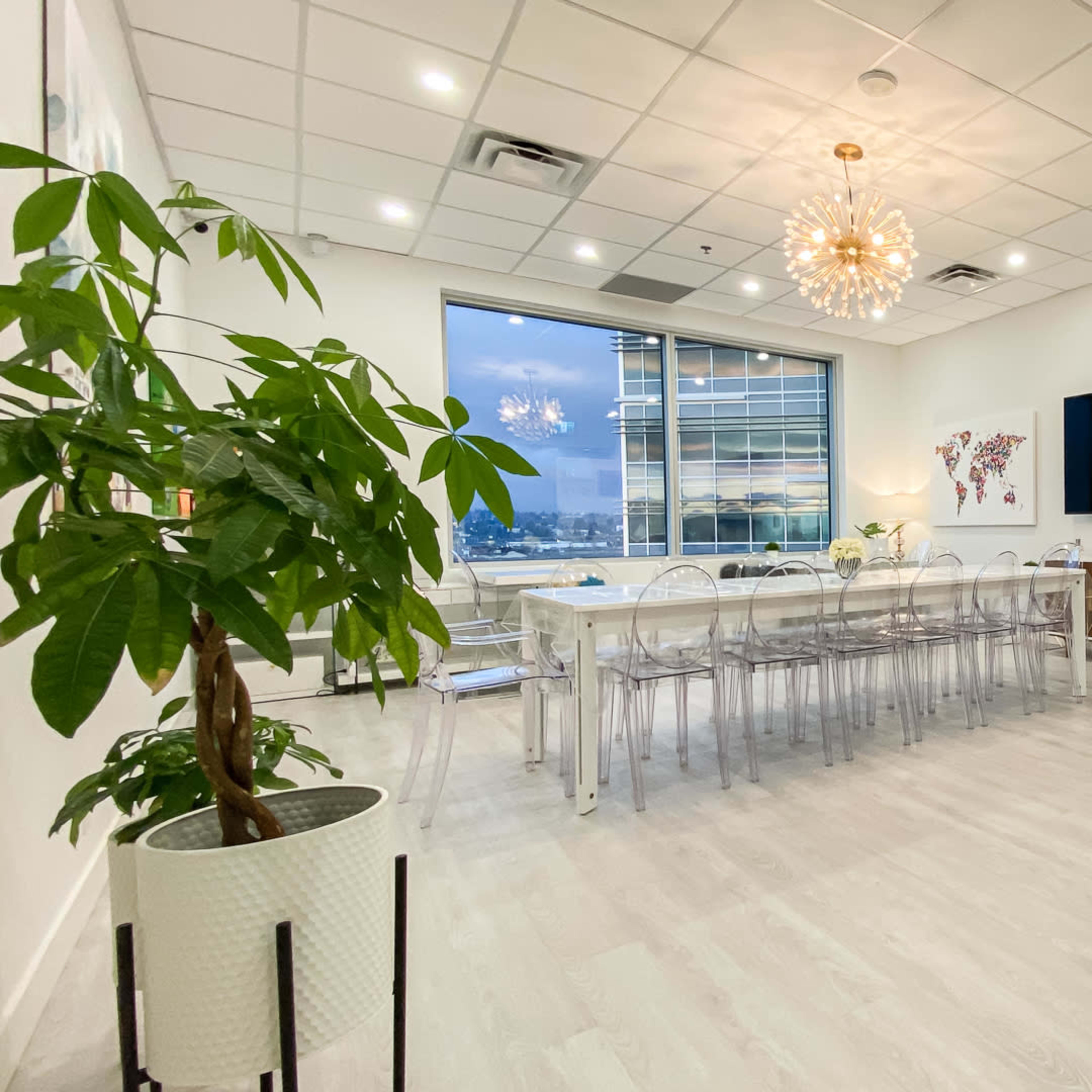 A modern conference room with a long table, transparent chairs, a large TV, and a view of the city through a window.