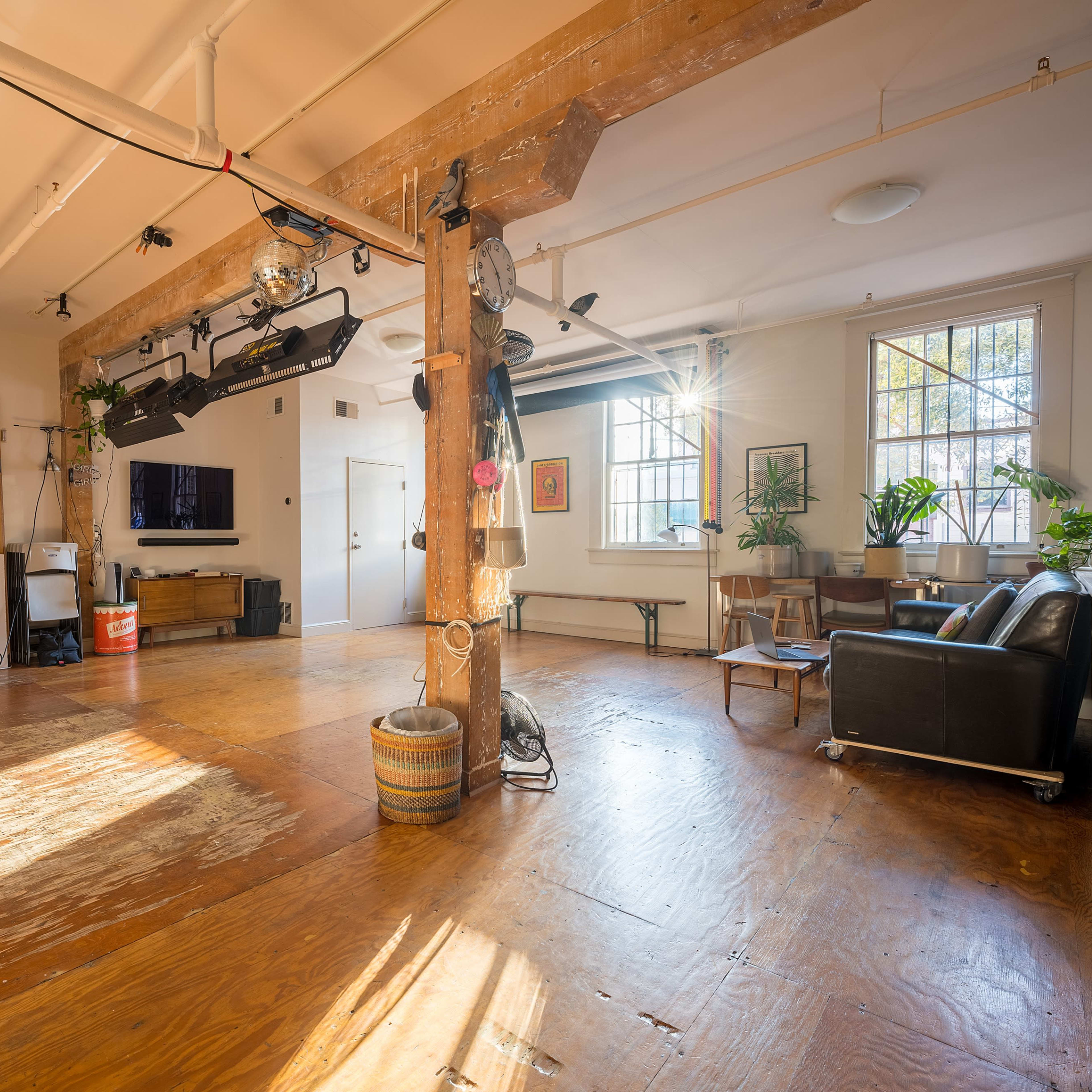 A spacious, well-lit studio interior with wooden floors, large windows, and various furniture and equipment arranged throughout the space.