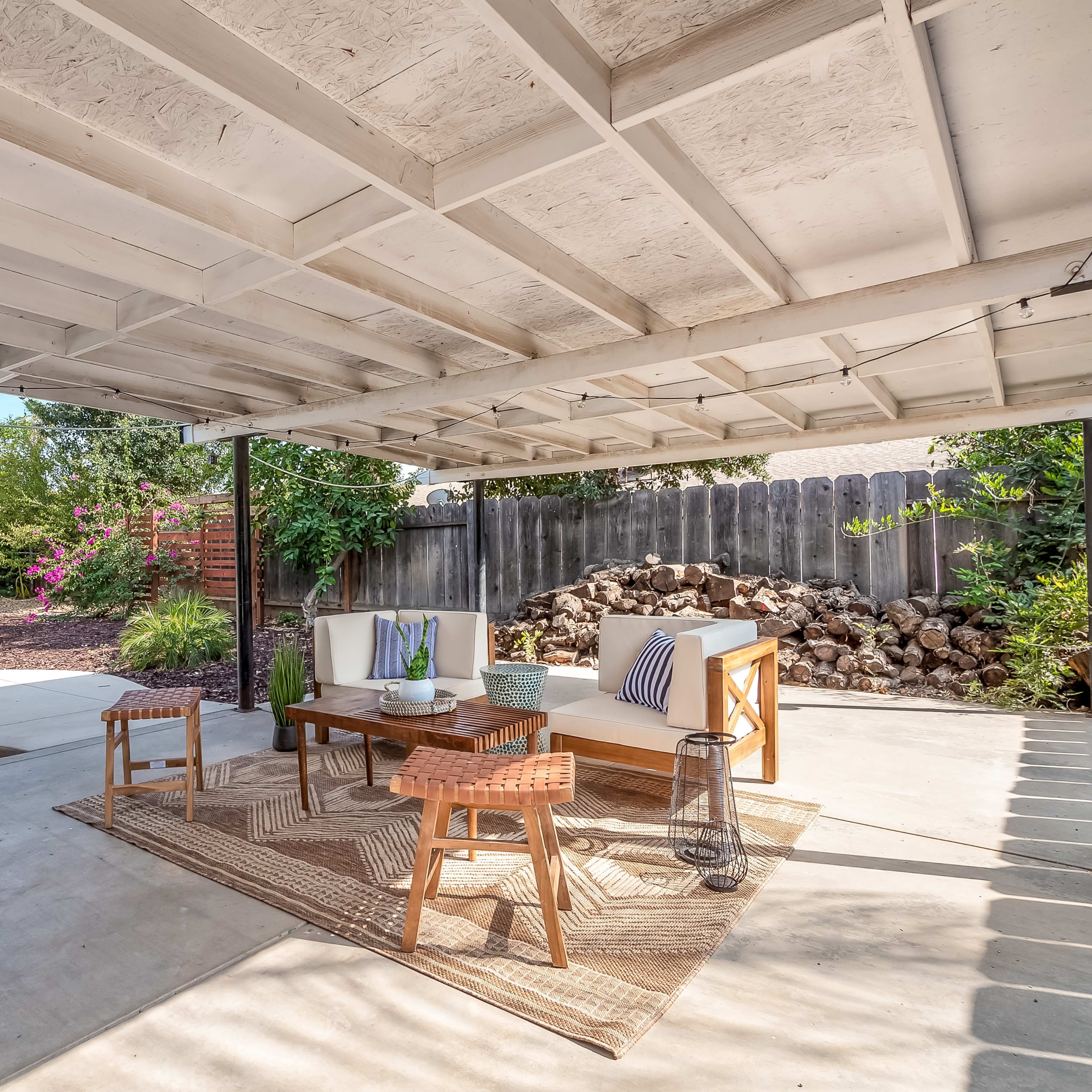 A covered outdoor seating area with a table and chairs surrounded by greenery and a stone wall.