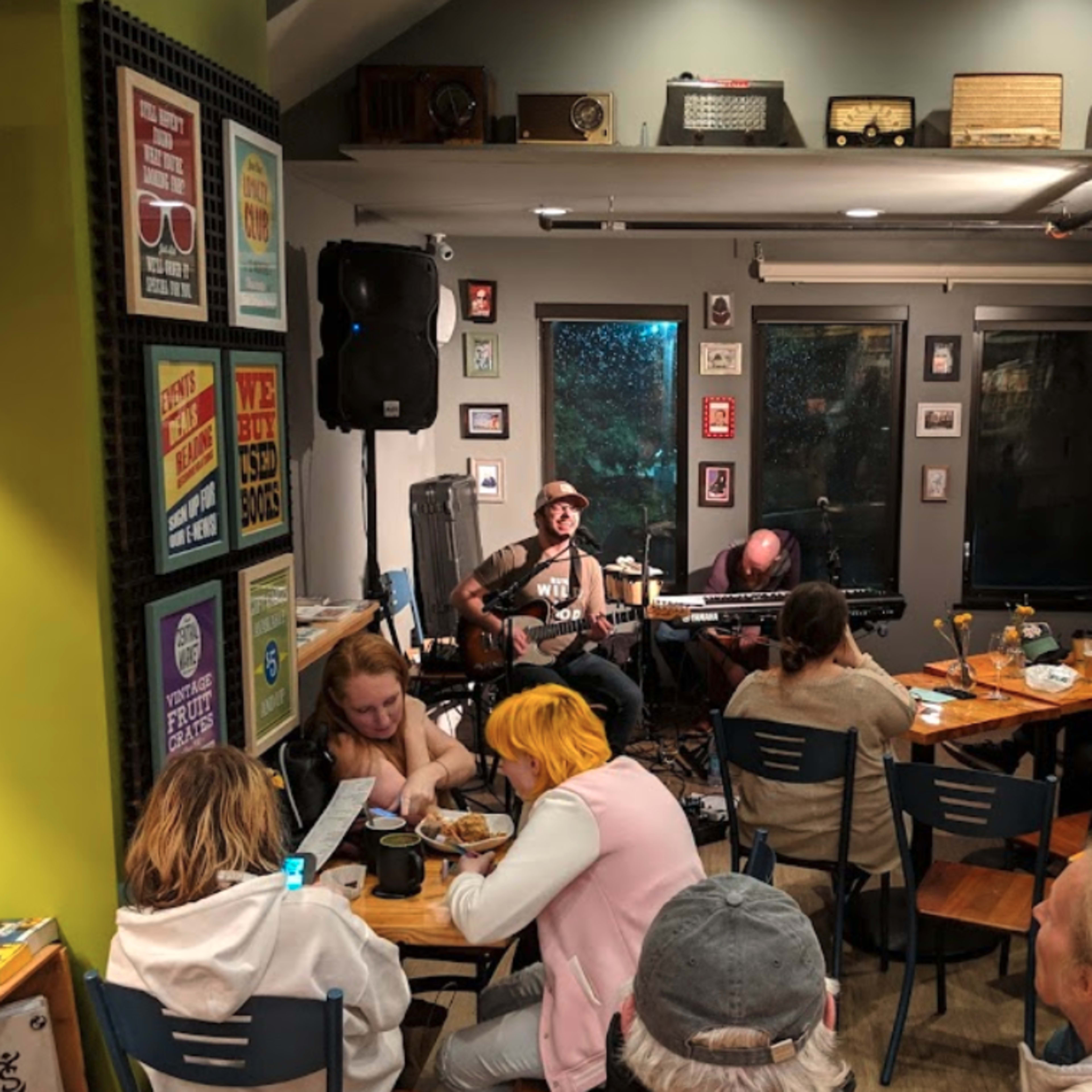 A small crowd listens to a musician performing with a guitar in a cozy cafe decorated with vintage items and framed posters.