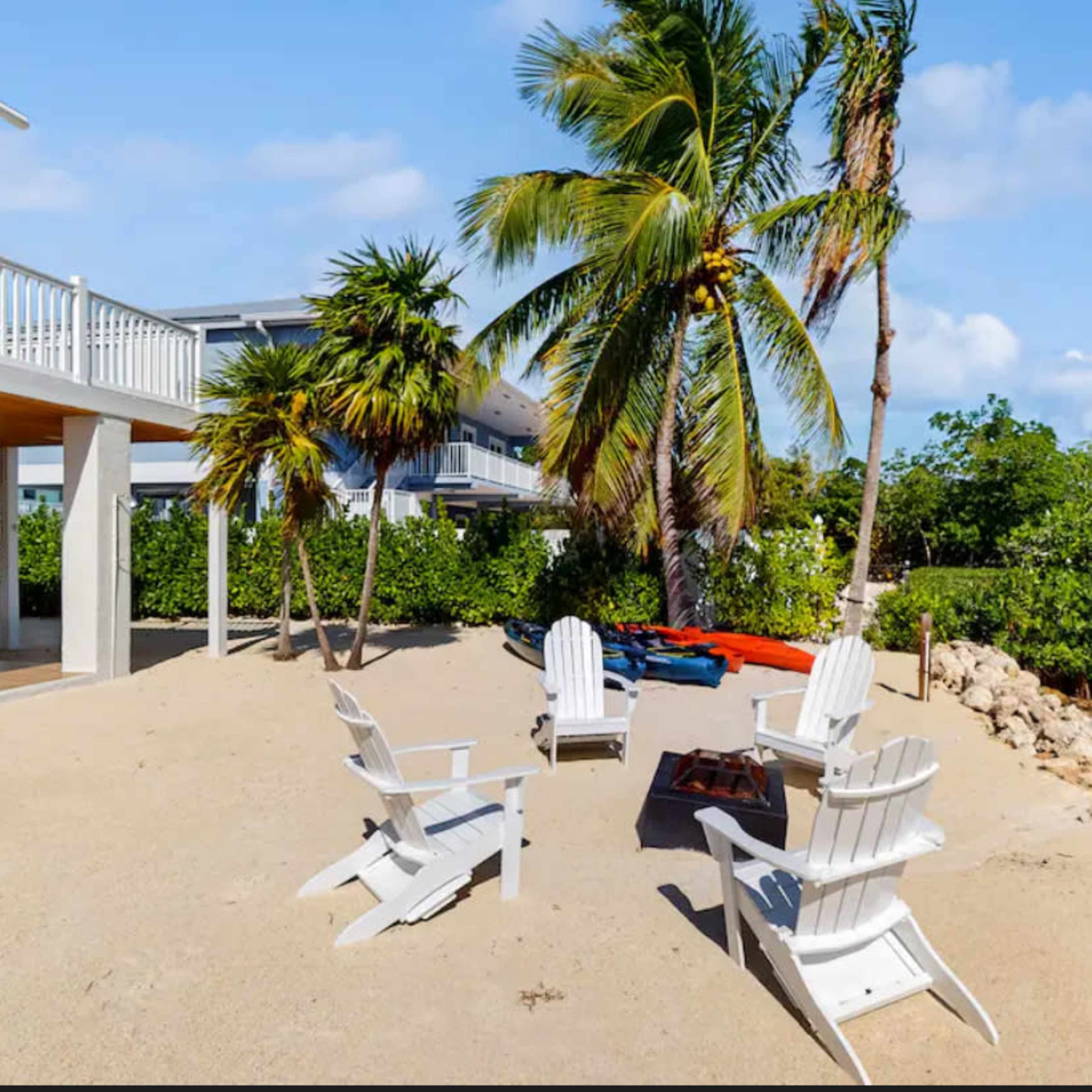A sandy beach area with four white Adirondack chairs arranged around a small fire pit, set against a backdrop of palm trees and a waterfront property.