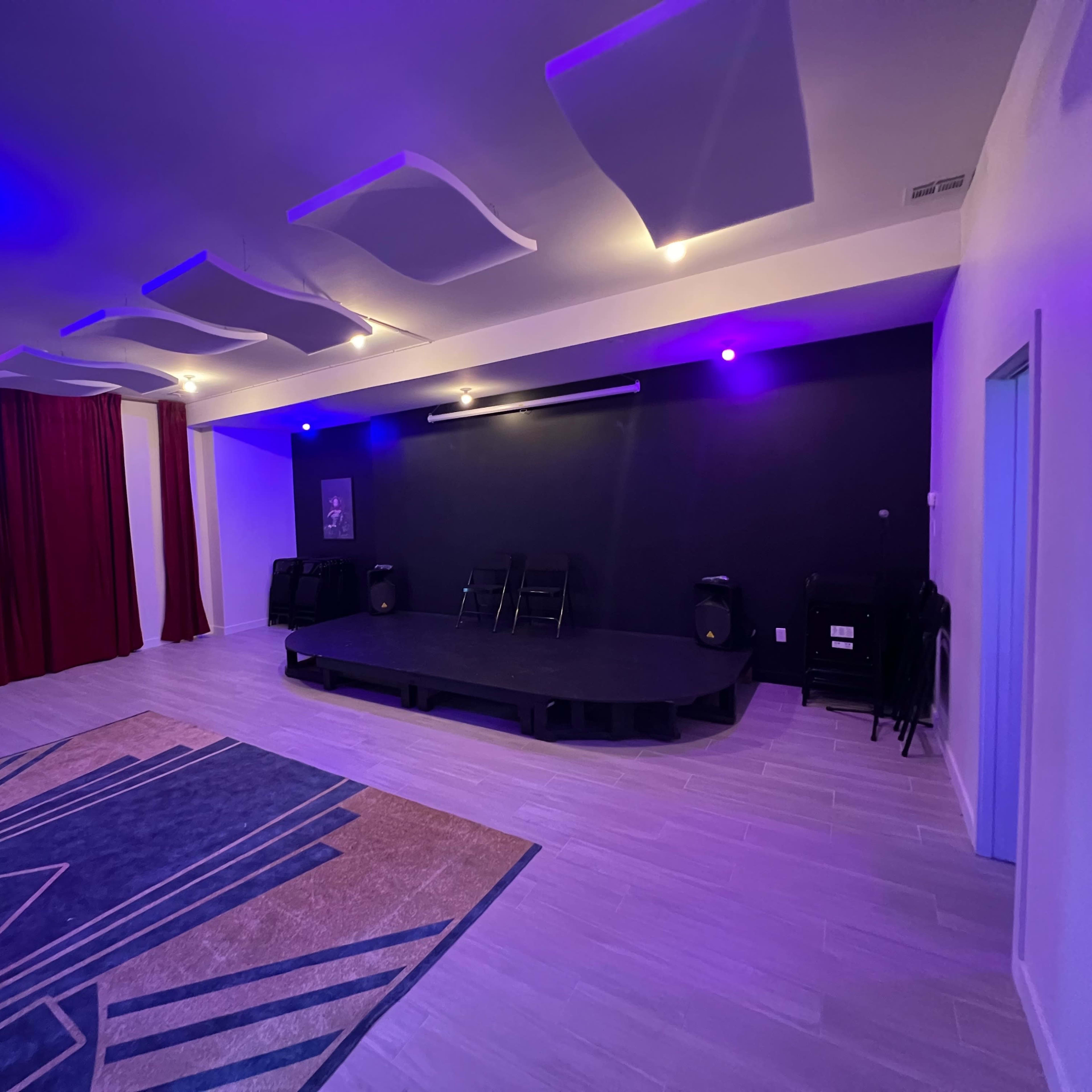 A spacious room with a stage, purple lighting, and sound equipment, featuring a red curtain on one side and a patterned carpet on the floor.