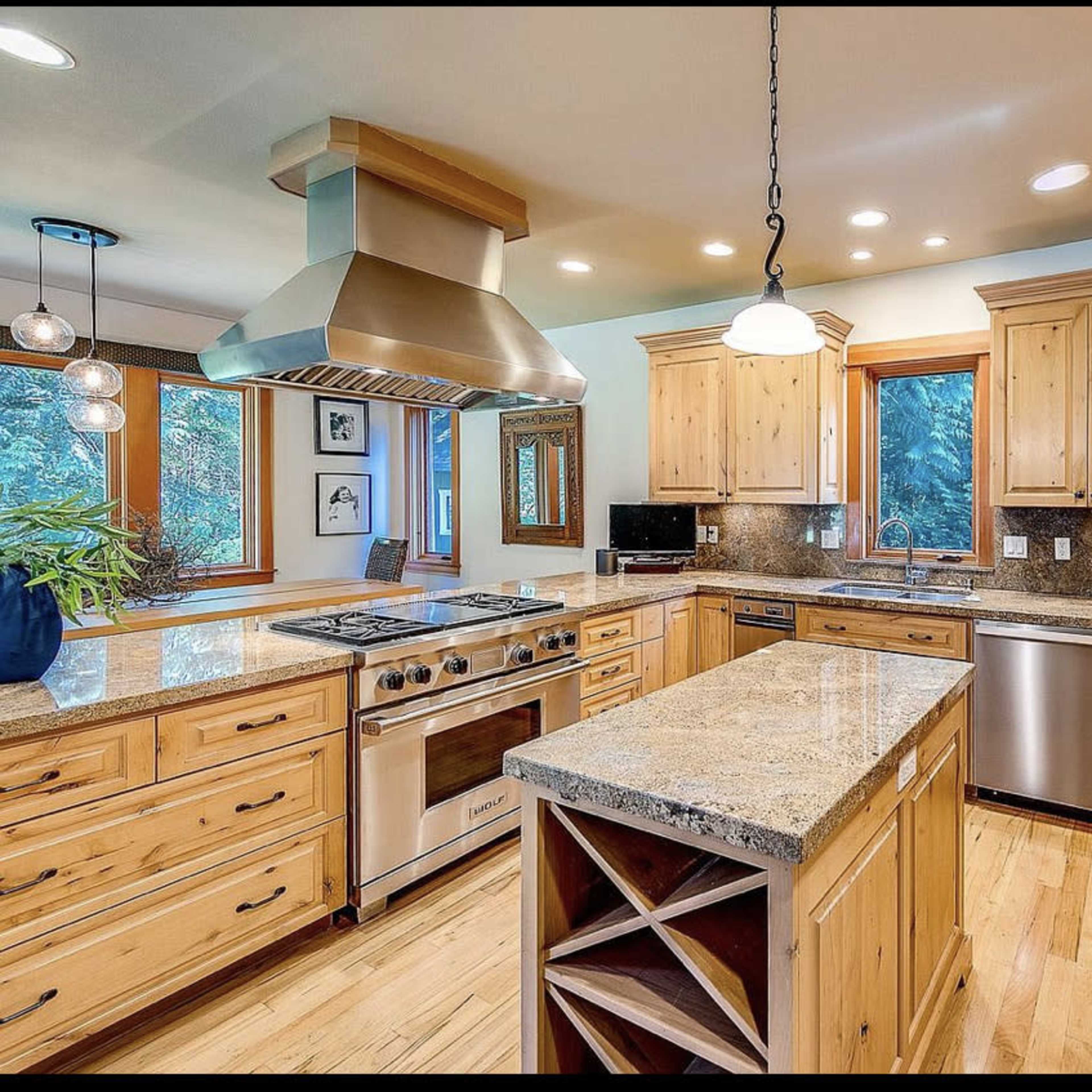 The kitchen features wooden cabinetry, a large island with a wine rack, and stainless steel appliances beneath a hooded range.