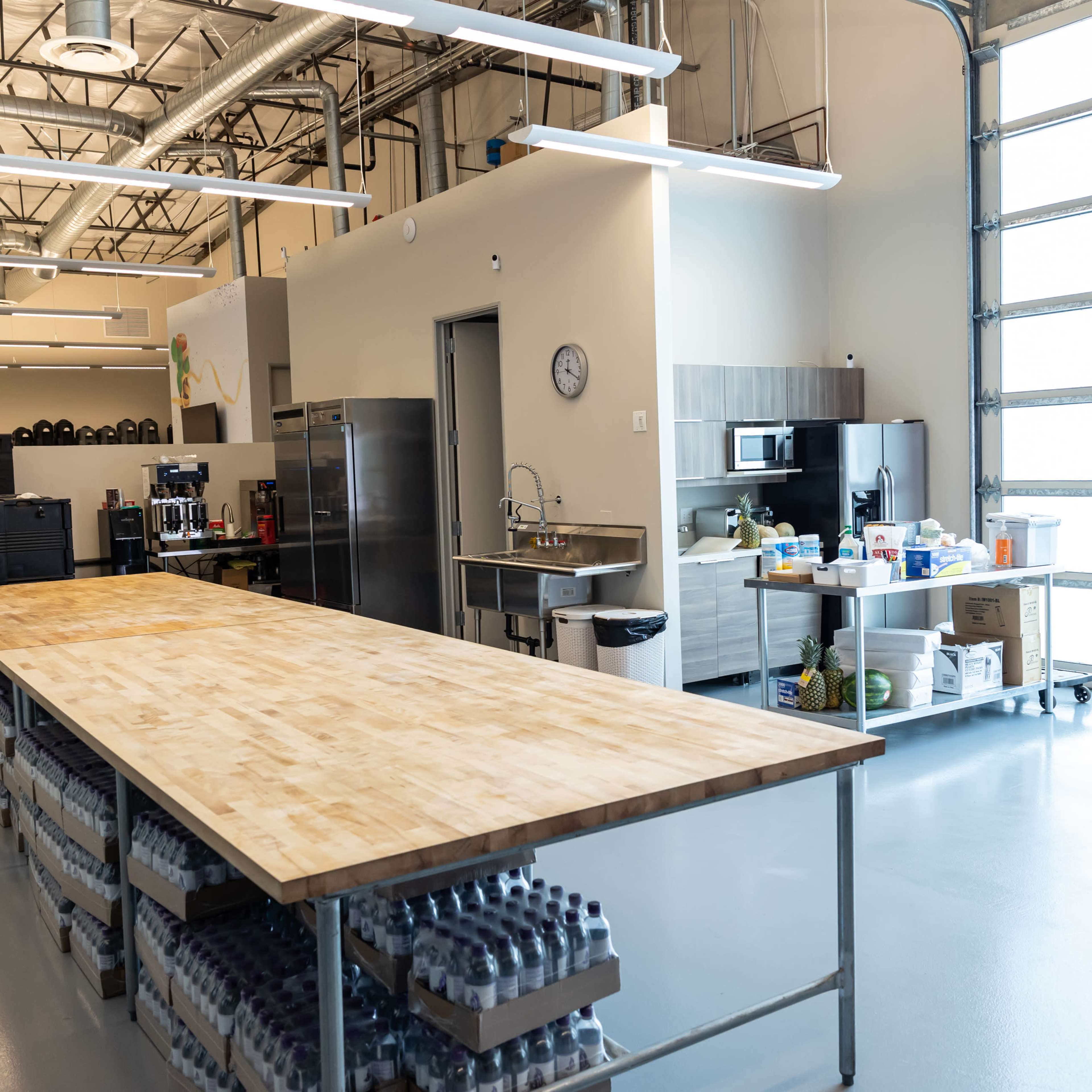 A spacious kitchen area with a large wooden table, a stainless steel kitchen setup, and storage shelves filled with supplies.