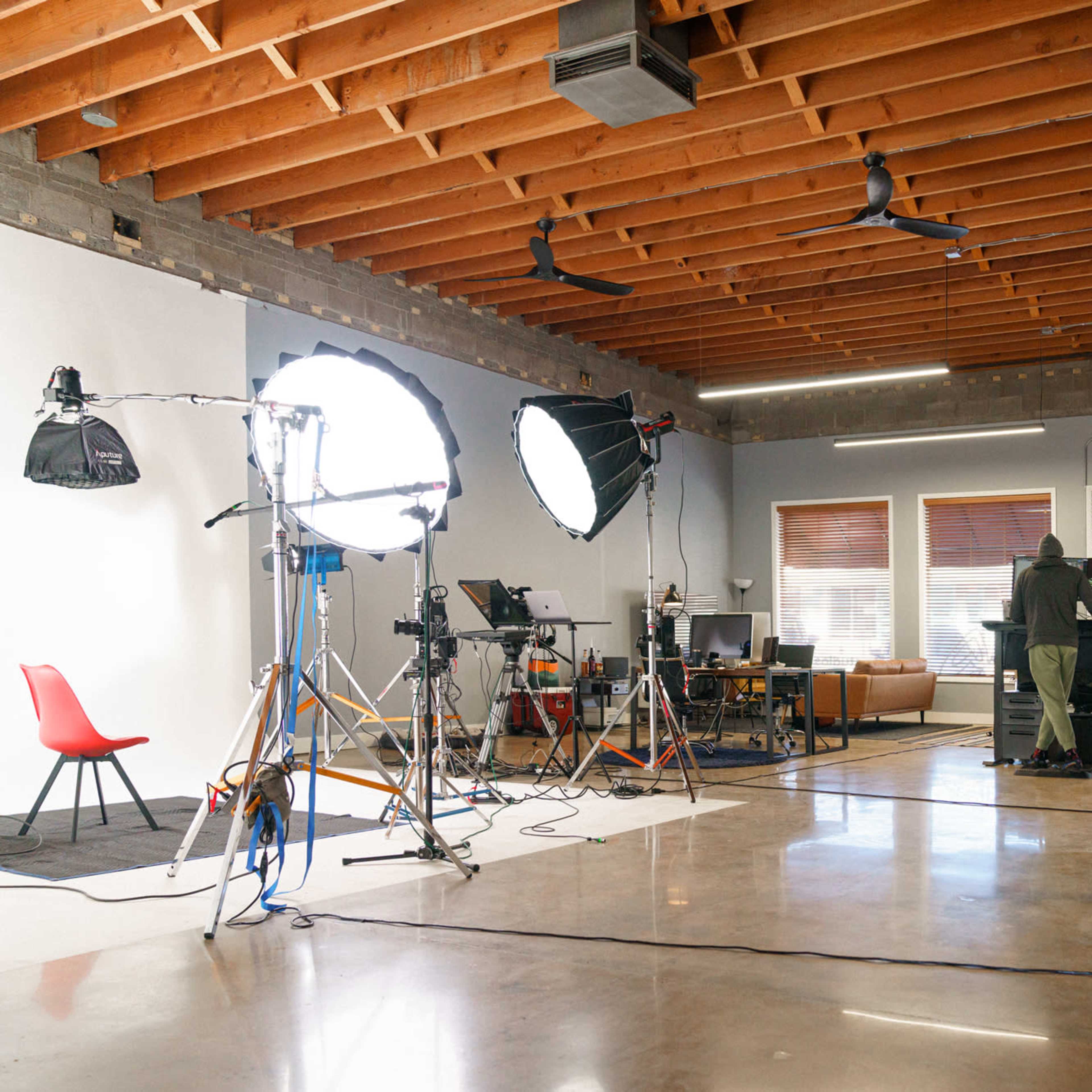 A spacious studio with various lighting setups and equipment, featuring a person working at a desk in the background.
