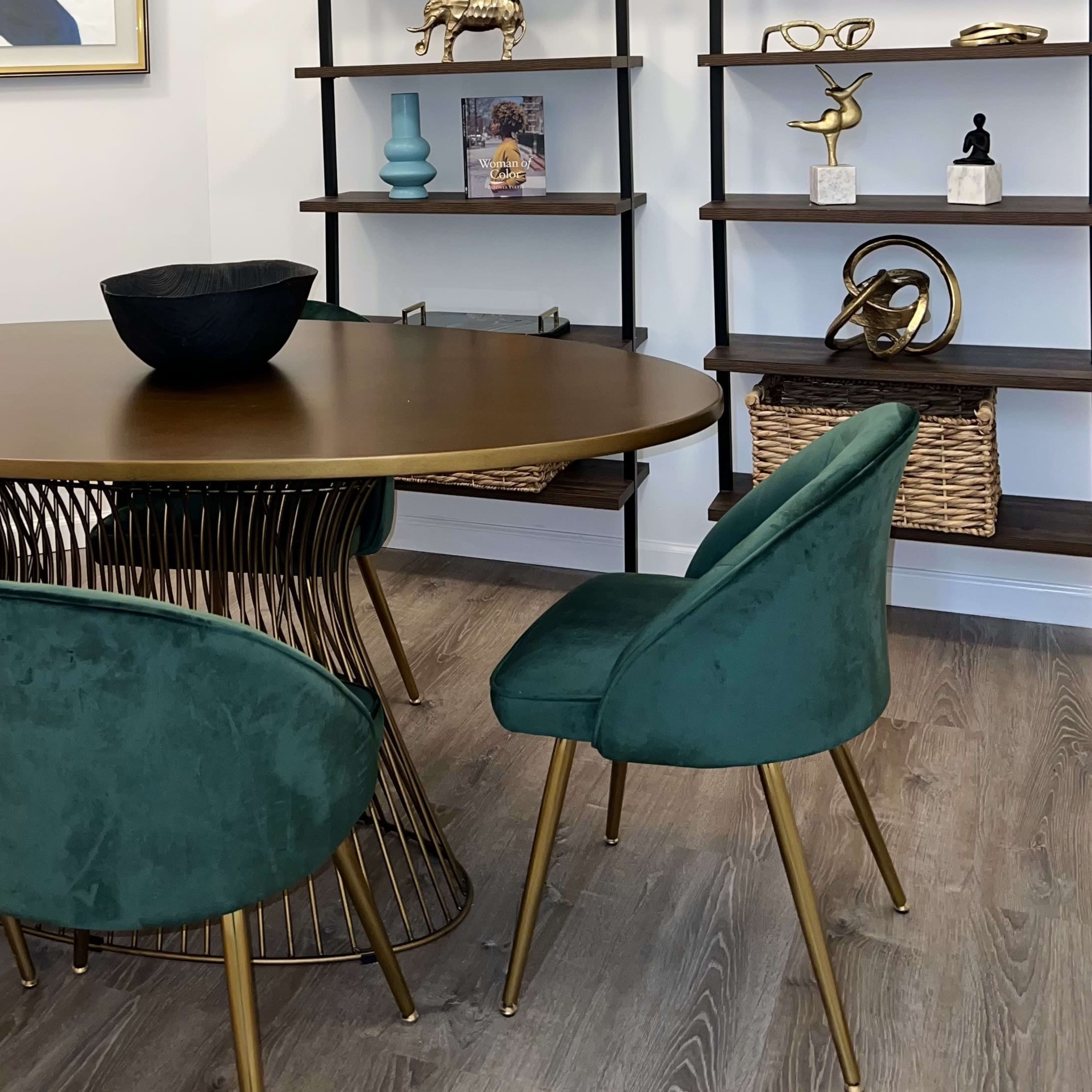 A round table with a metal base is surrounded by four green velvet chairs in a well-decorated room featuring shelves with various decorative items.