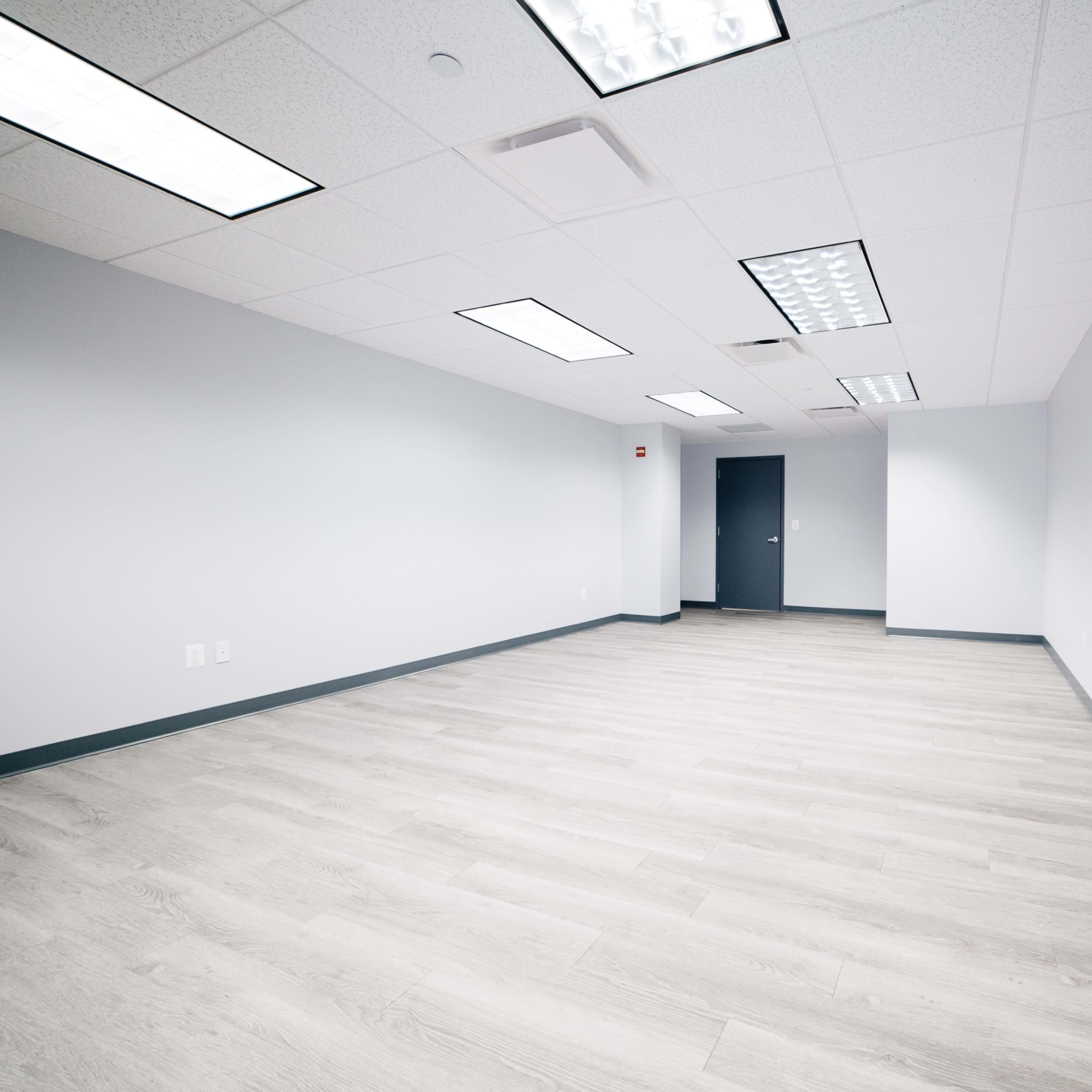 An empty office space with light-colored flooring and gray walls, featuring a single door on one side.