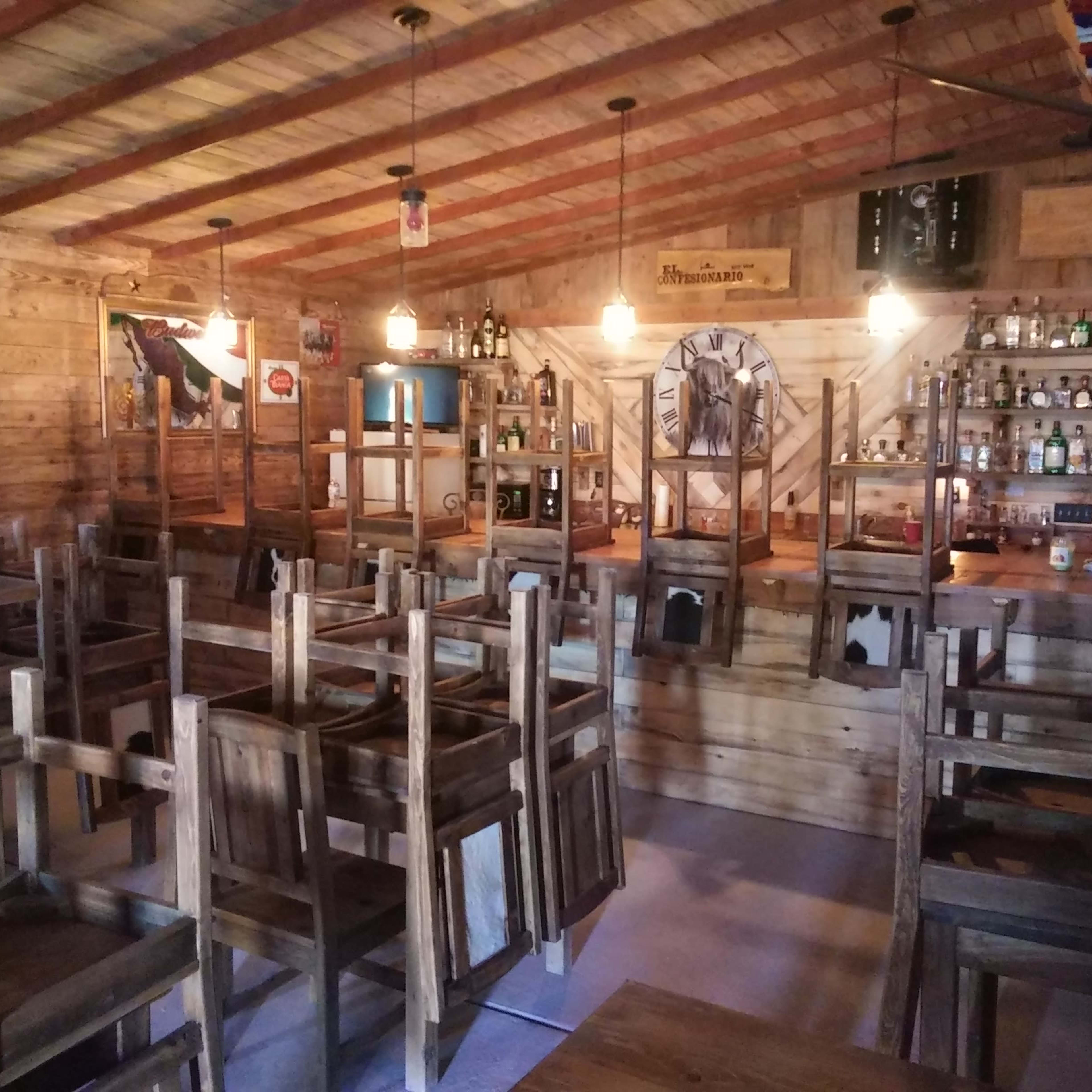 A rustic bar interior with wooden chairs stacked on tables and a counter stocked with bottles in the background.