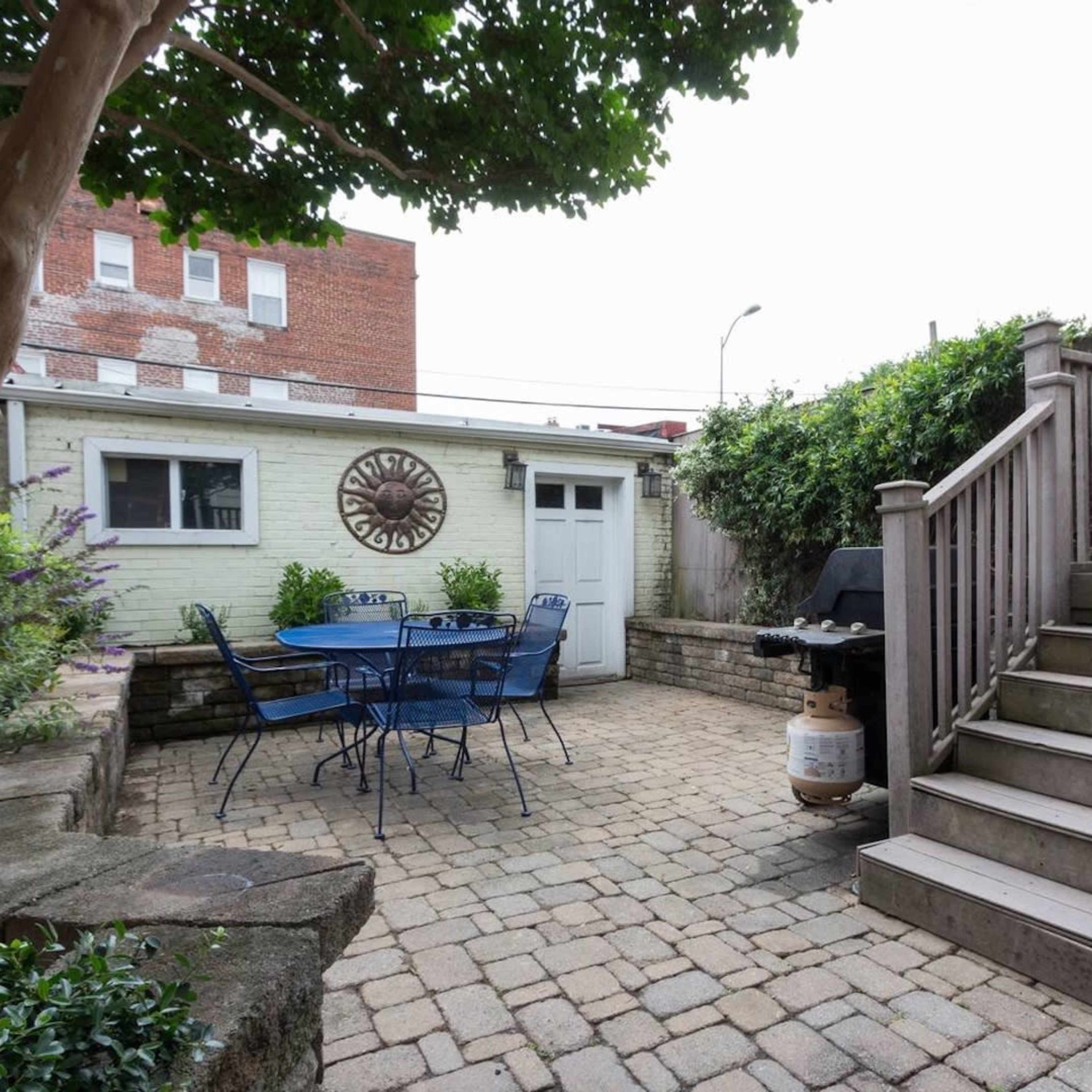 A paved backyard area features a table and chairs, a grill, and stairs leading to an elevated deck.