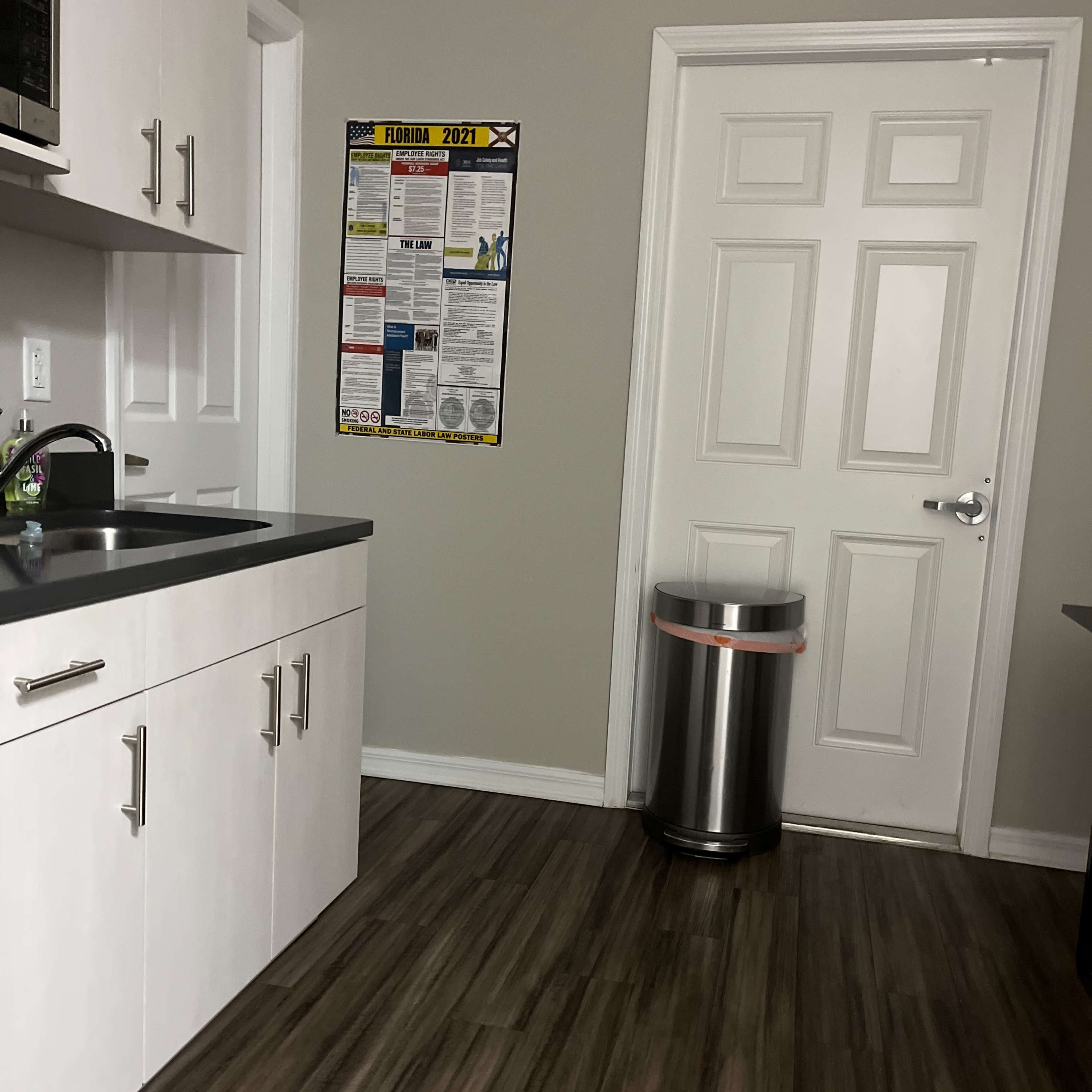A kitchen corner features a sink and countertop, a poster on the wall, and a trash can beside a closed door.
