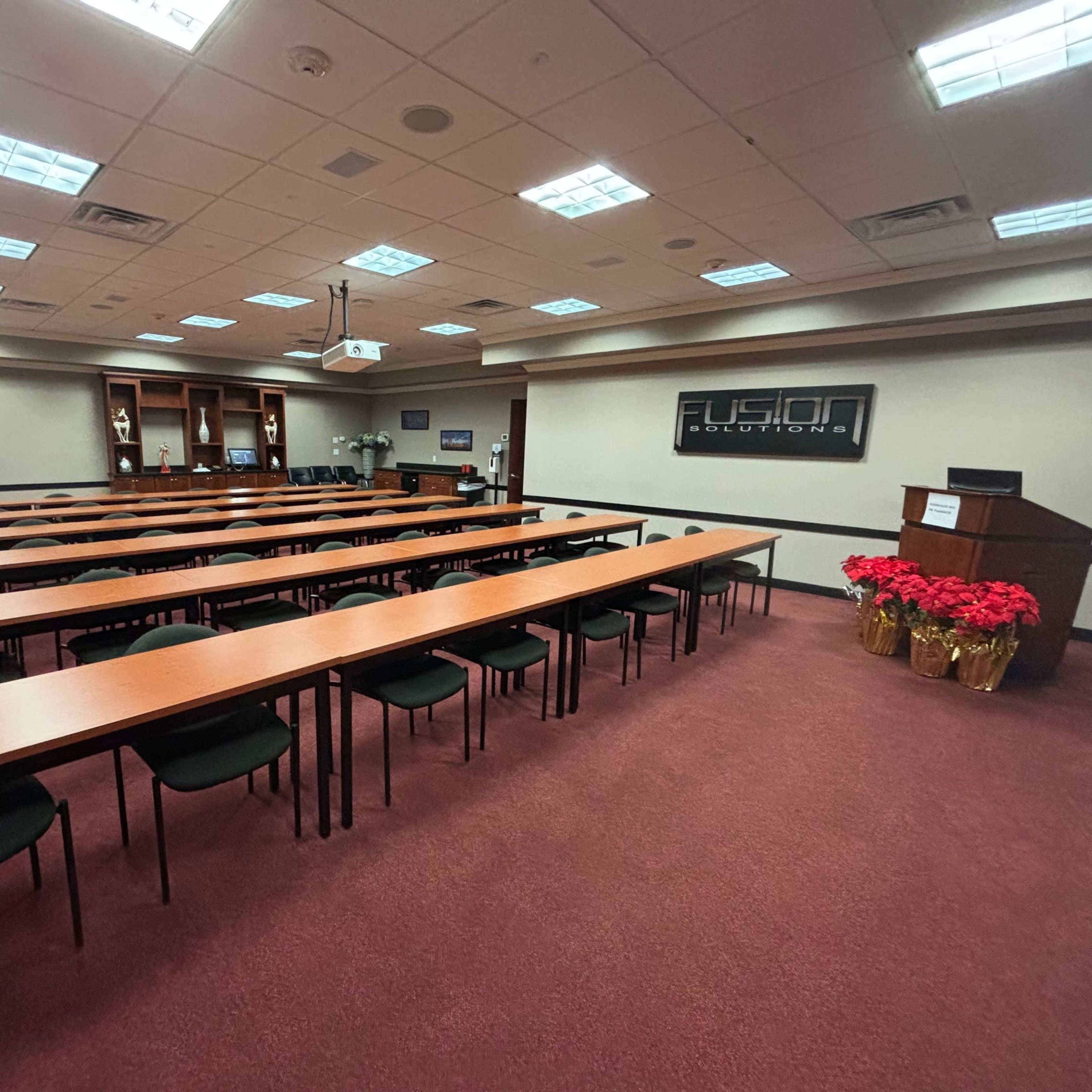 A conference room arranged with multiple long tables and chairs, featuring a podium at one end and a display screen on the wall.