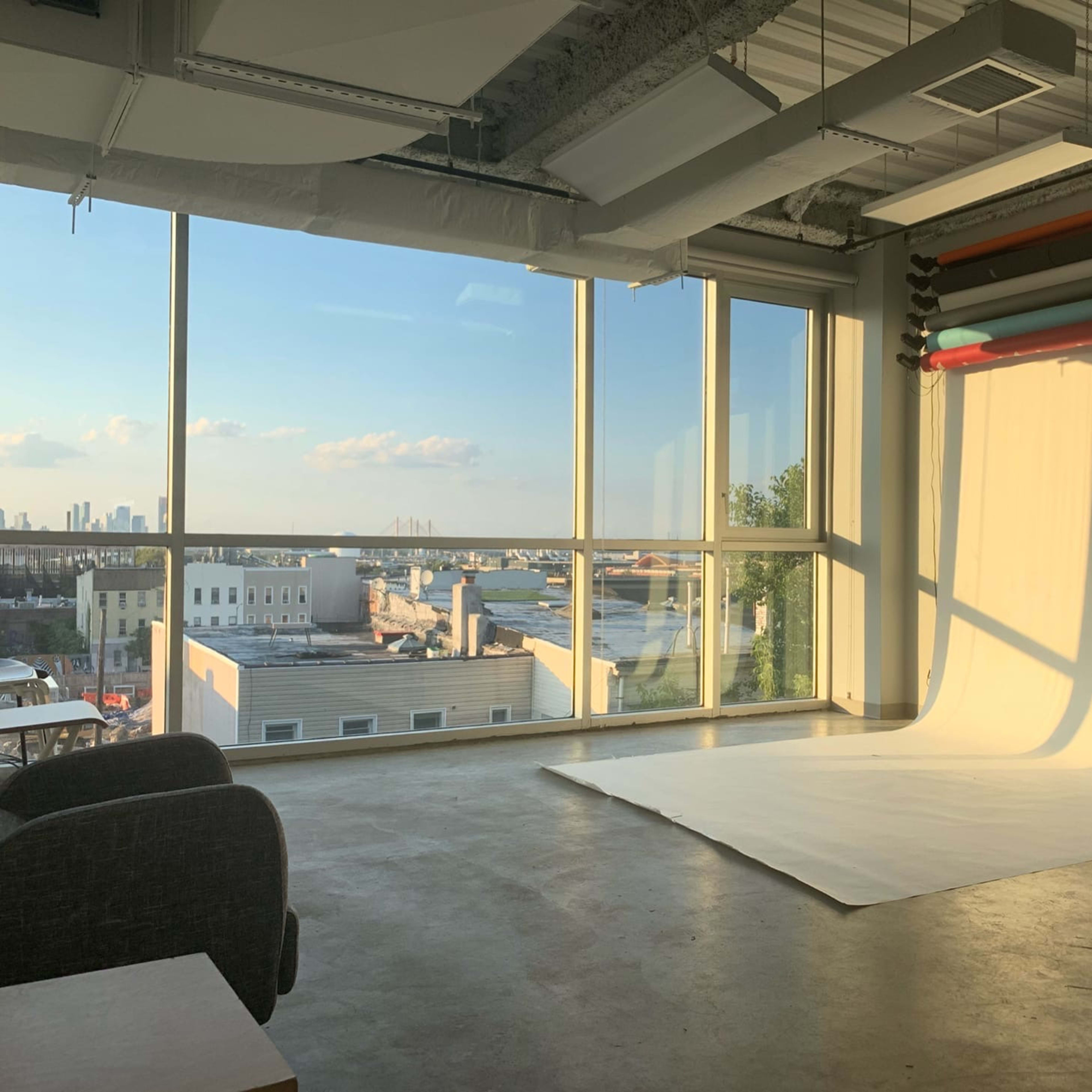 A photography studio with a large window overlooks a cityscape, featuring a white backdrop and colored rolls hanging on the wall.