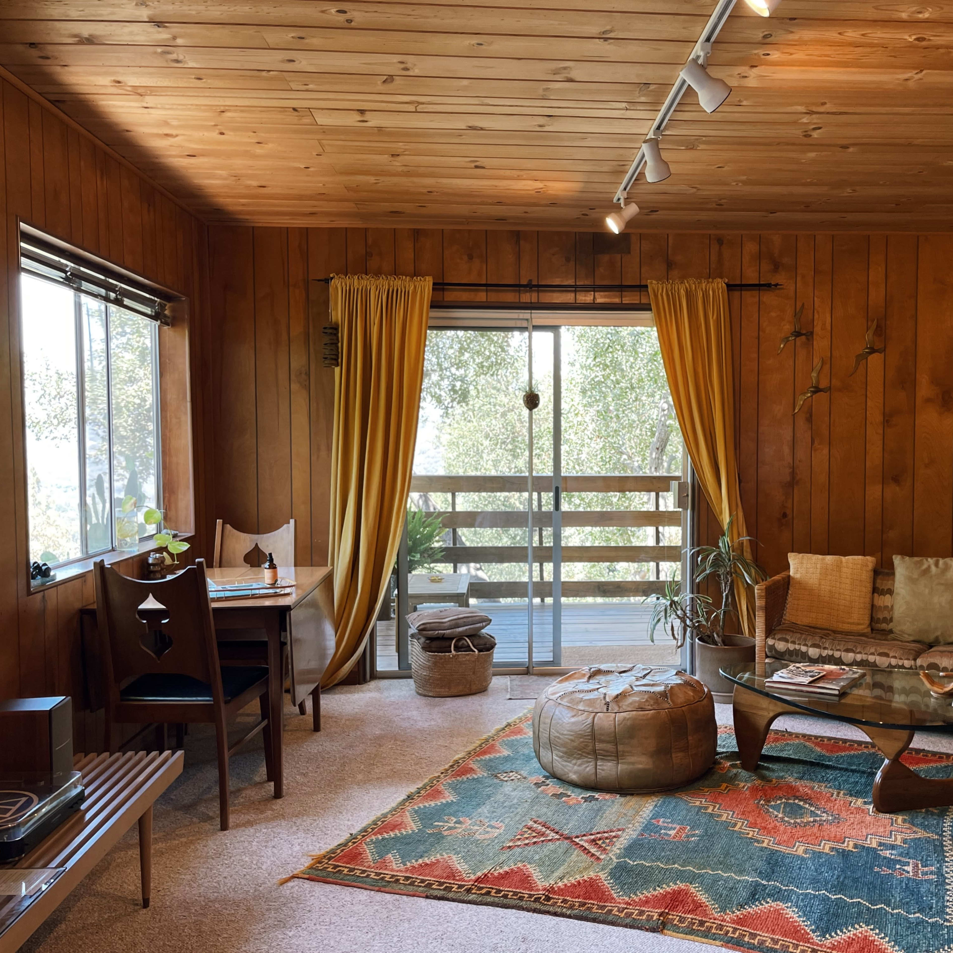 A cozy living room with wooden walls, a patterned rug, a sofa, and large windows leading to a view outside.