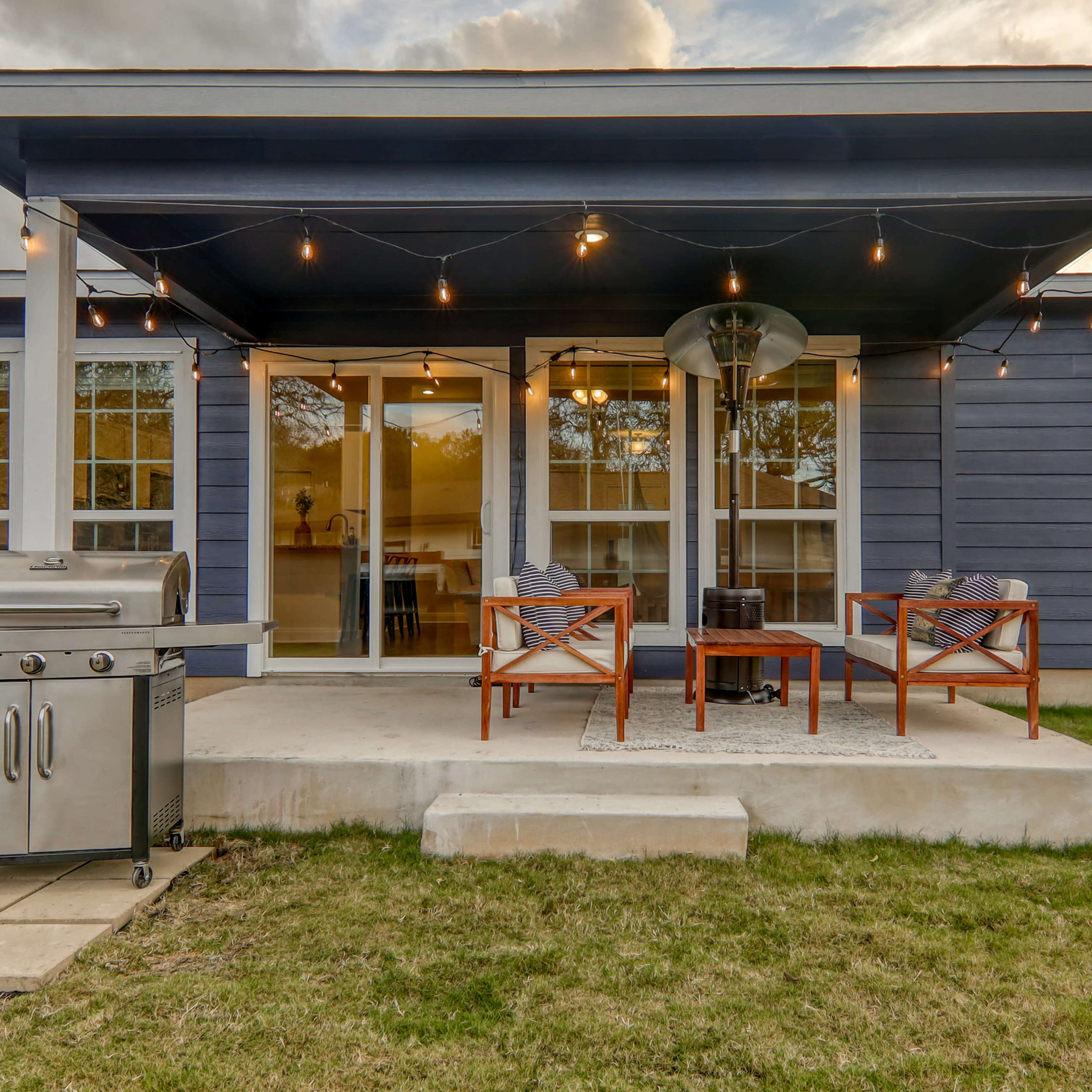 A patio with two chairs and a table, alongside a grill, beneath a covered porch with string lights.