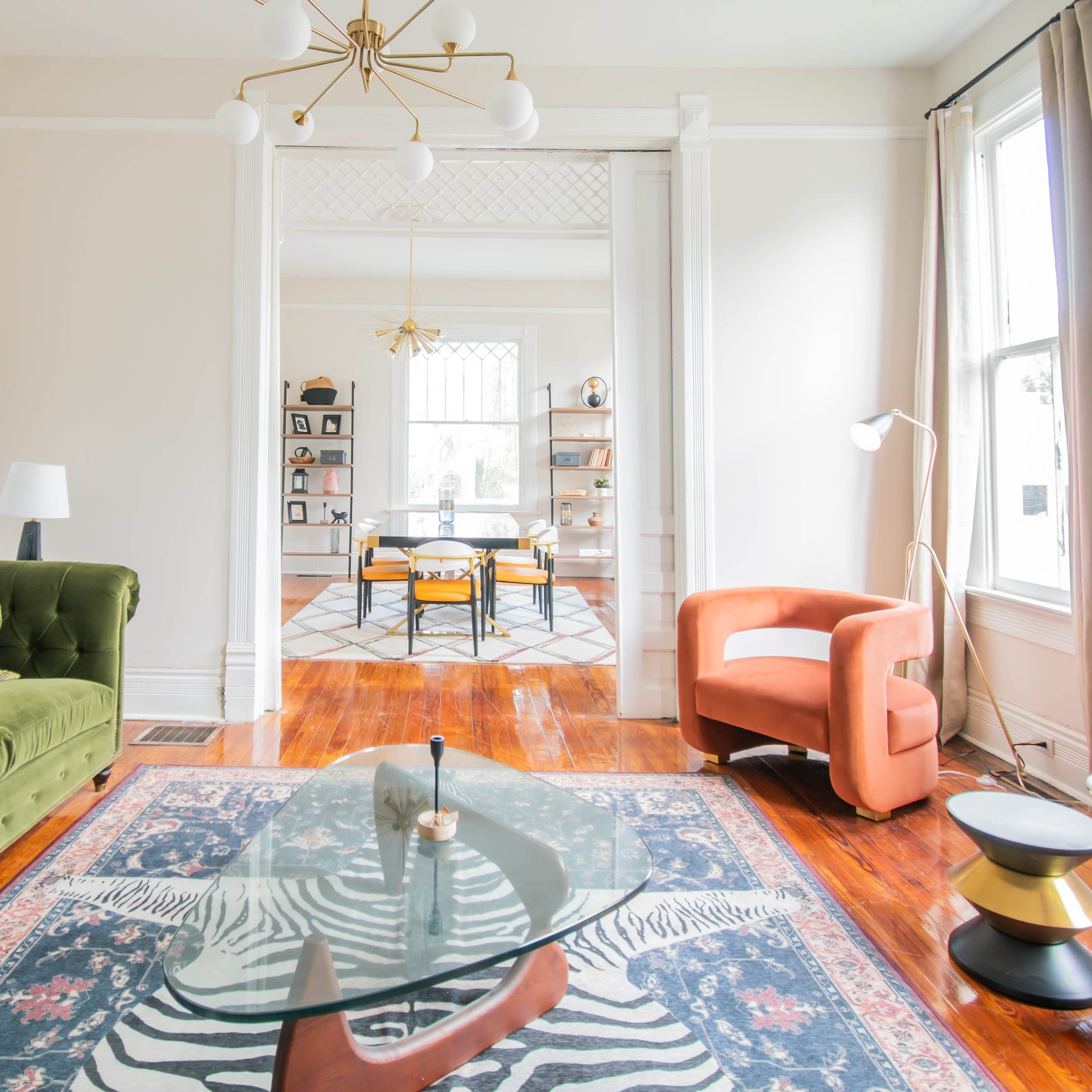 A green velvet sofa and an orange armchair are arranged in a bright room with hardwood floors and large windows, featuring a glass coffee table and a black and white zebra-patterned rug.