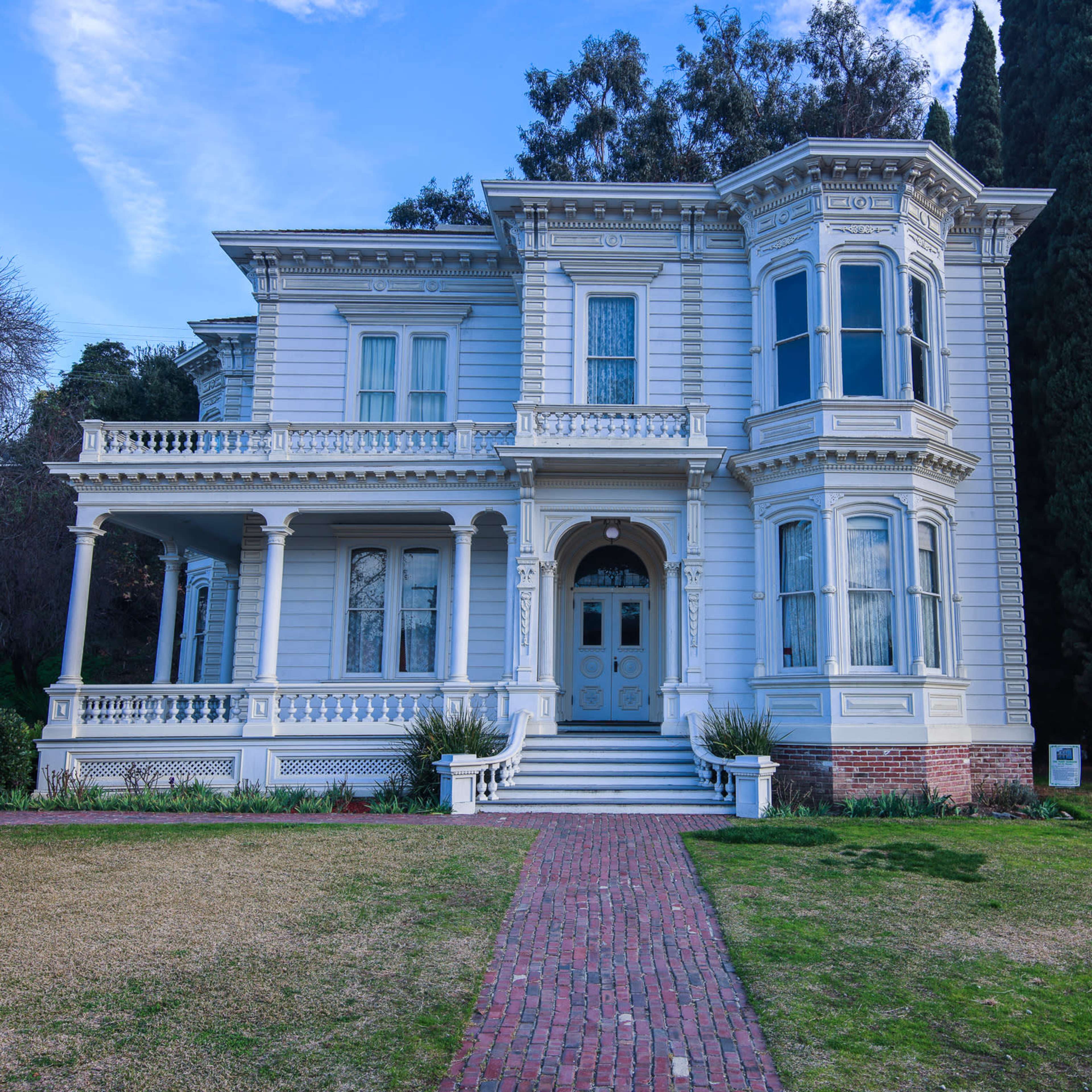 A large, ornate Victorian-style house with a front porch, multiple windows, and a brick pathway leading to the entrance.