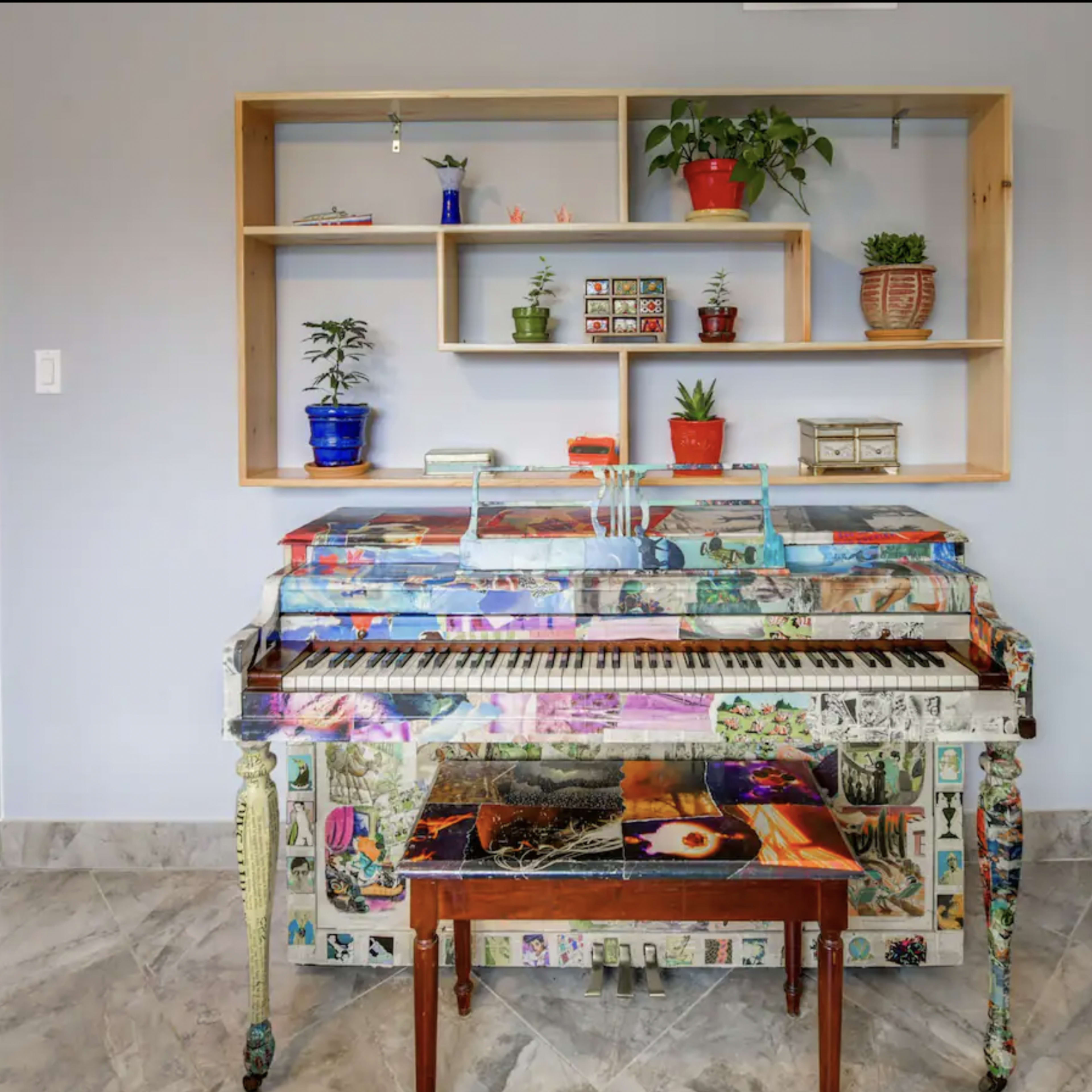 A decorated piano with colorful artwork and a small wooden table sits beneath a shelf containing various plants and decorative items against a plain wall.