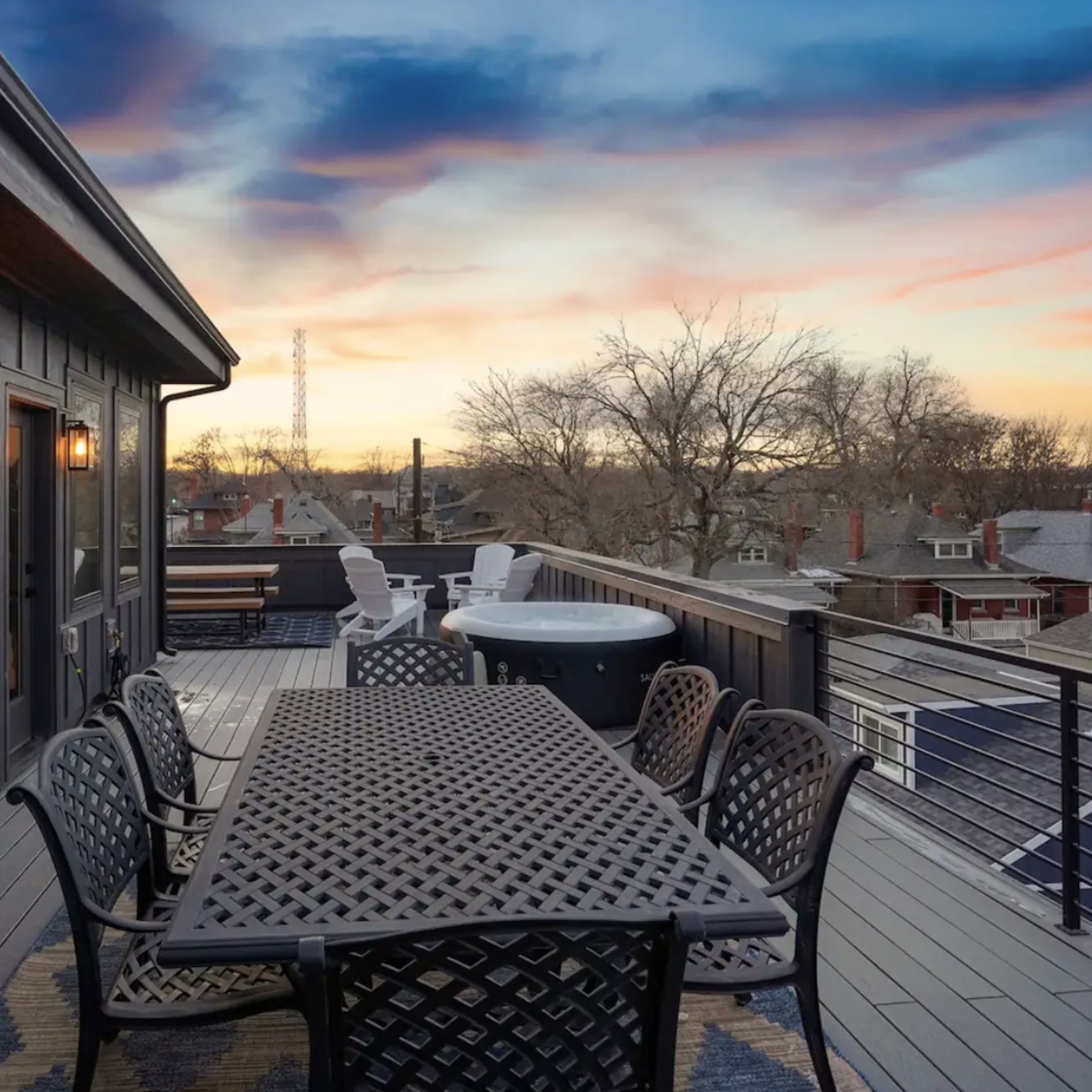 A rooftop terrace features a large table surrounded by chairs, with a hot tub nearby and a backdrop of a colorful sunset over the rooftops of nearby houses.