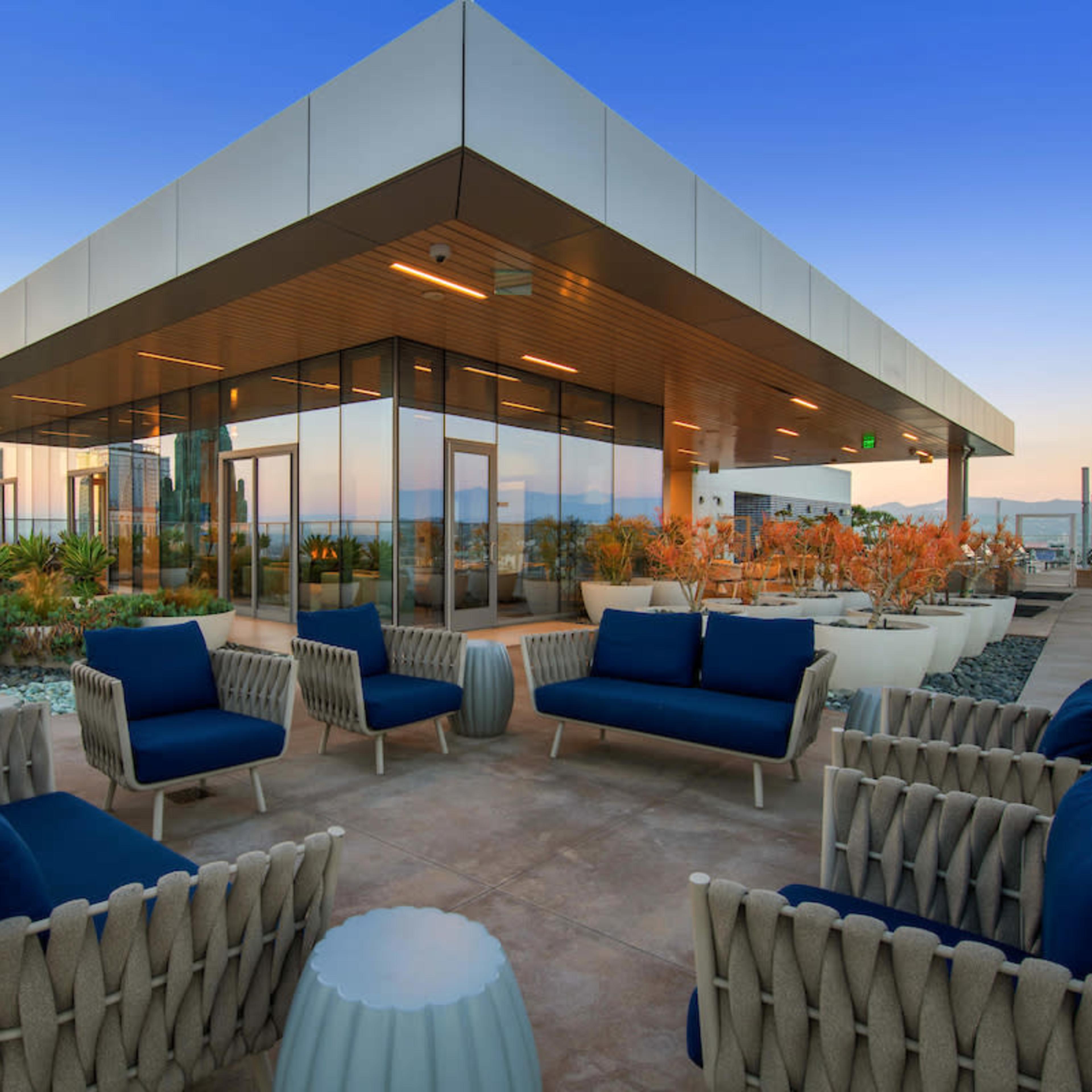 A modern rooftop lounge area with comfortable seating and planters, surrounded by the skyline at dusk.
