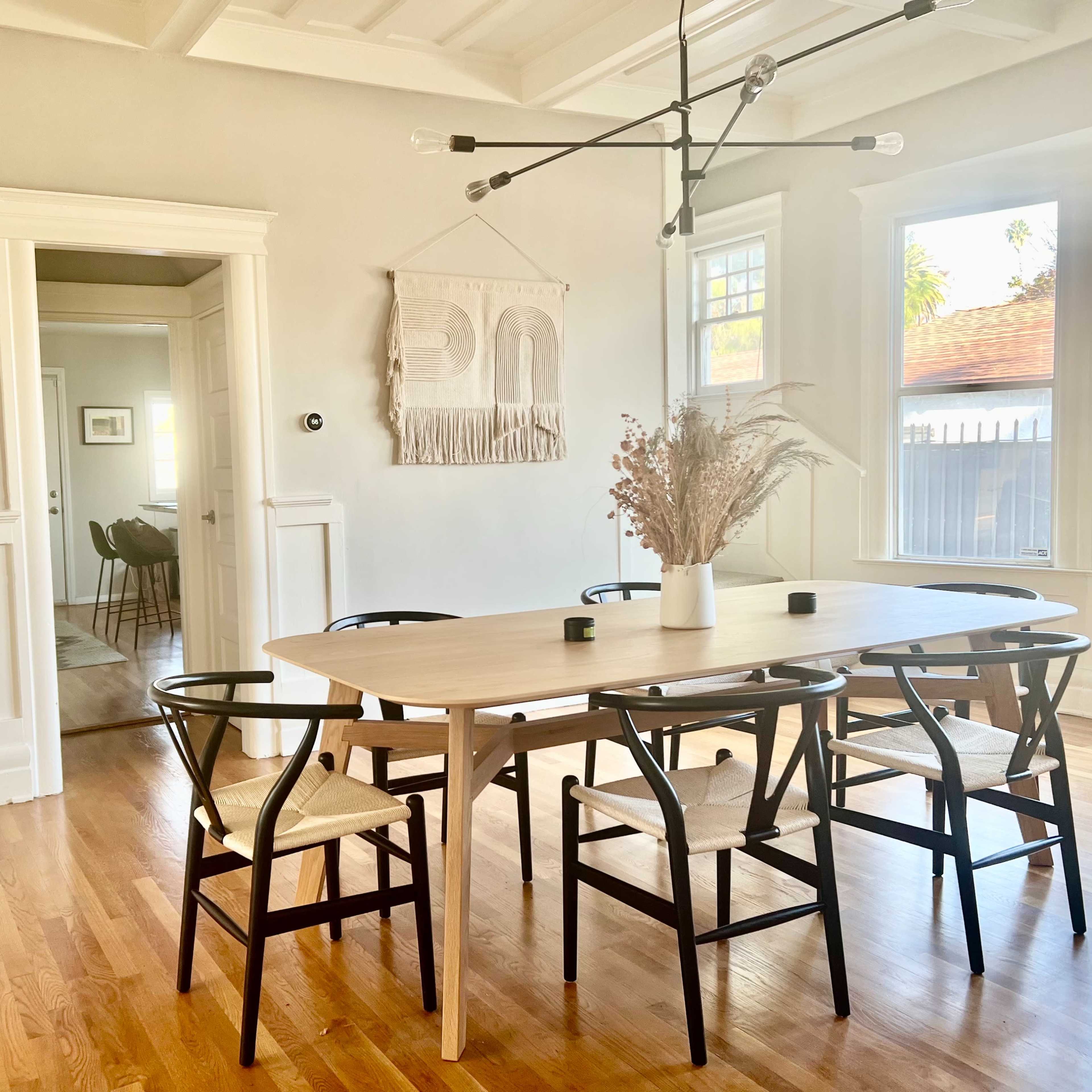 A light-filled dining room features a large table surrounded by six chairs and a decorative wall hanging.
