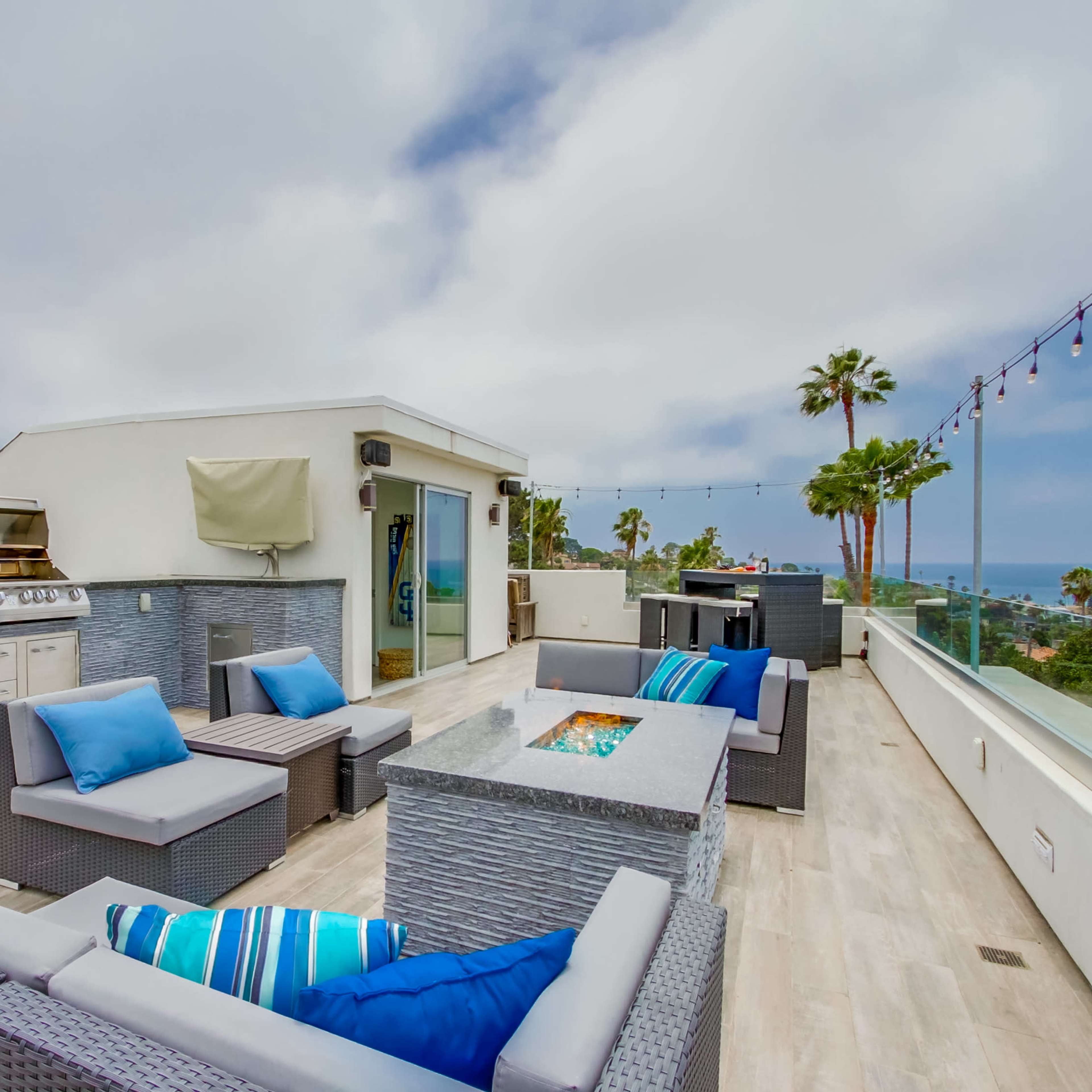 A rooftop terrace with modern outdoor furniture, a barbecue grill, and a view of the ocean under a cloudy sky.