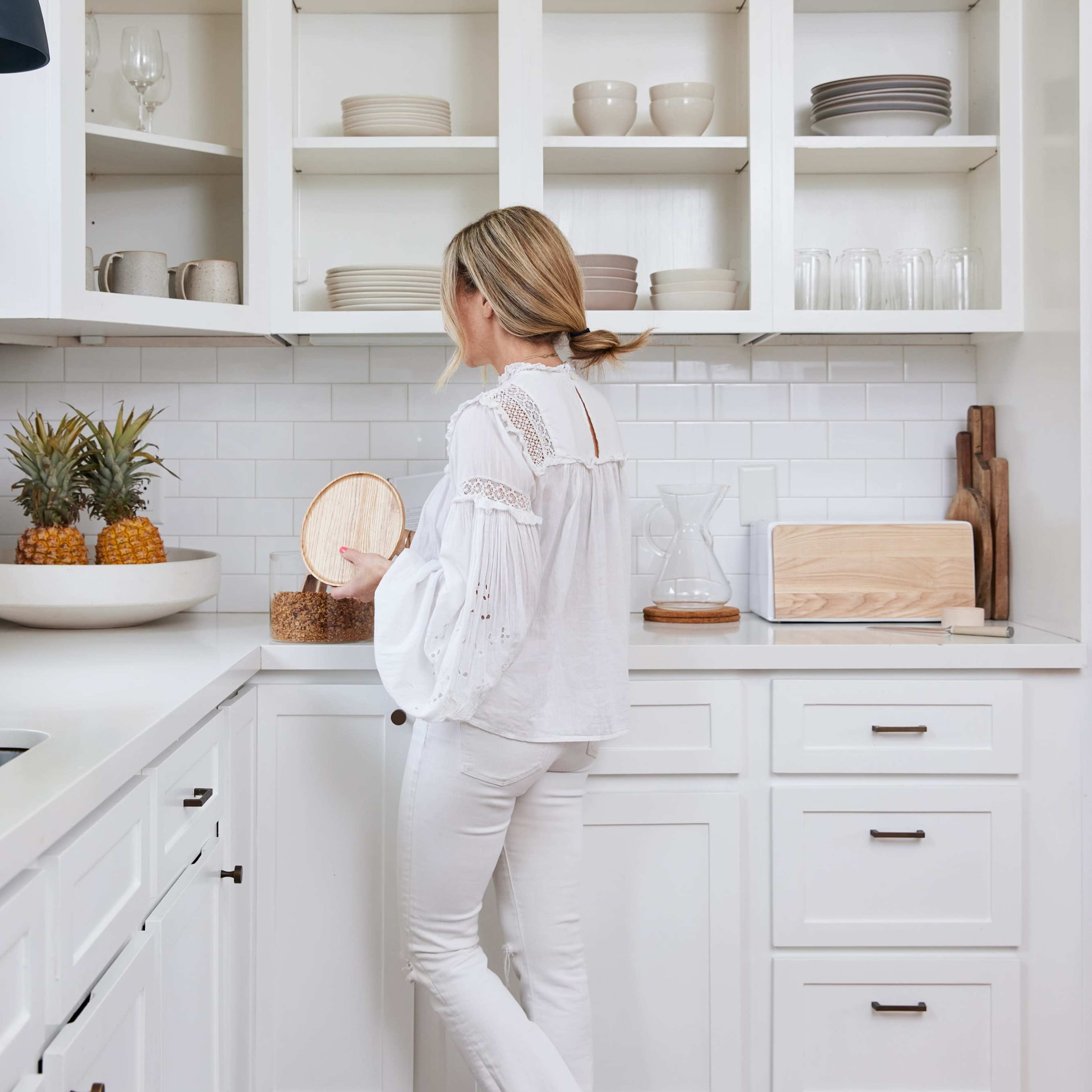 A person stands in a modern kitchen, inspecting items on a countertop next to an arrangement of pineapples.