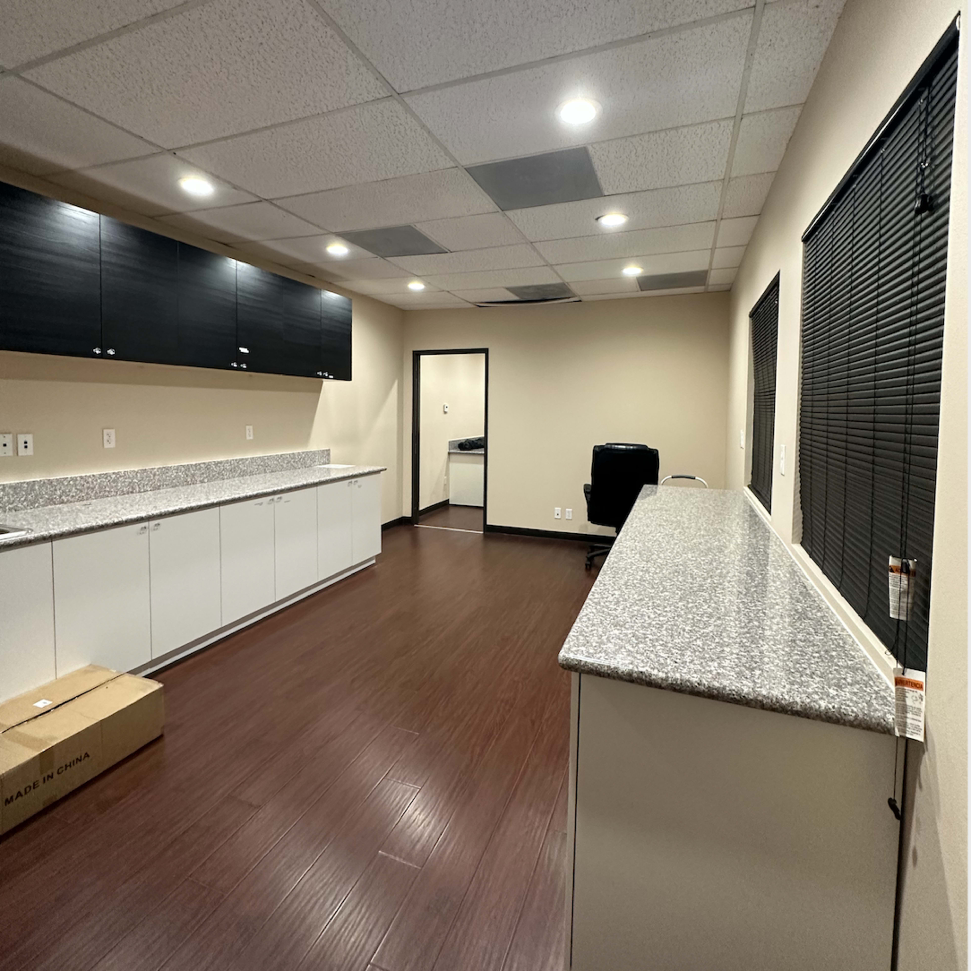 A modern office space features a streamlined design with a long countertop, dark cabinets on the wall, and hardwood flooring.