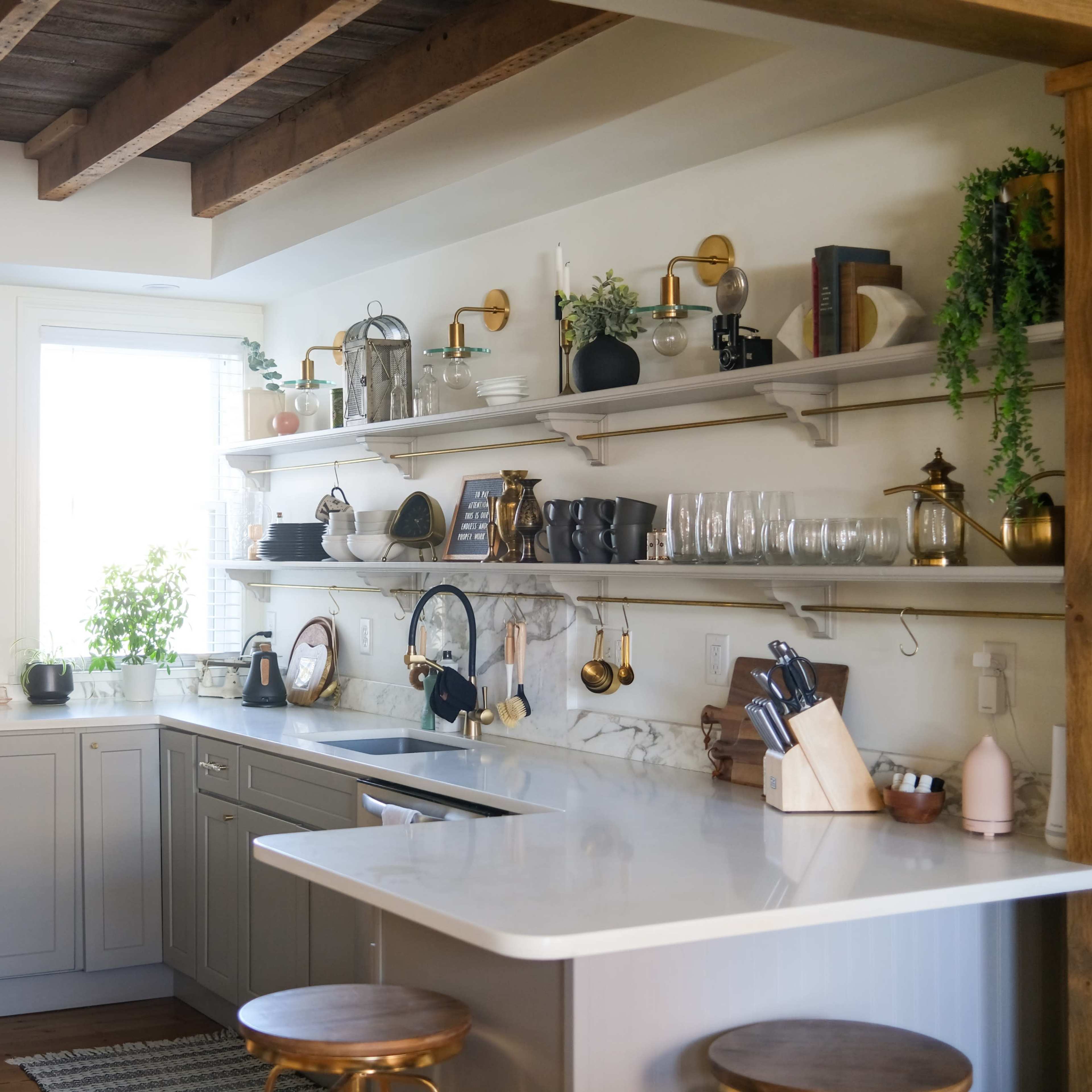 A modern kitchen with gray cabinets, a white countertop, open shelving displaying kitchenware, and wooden beams overhead.