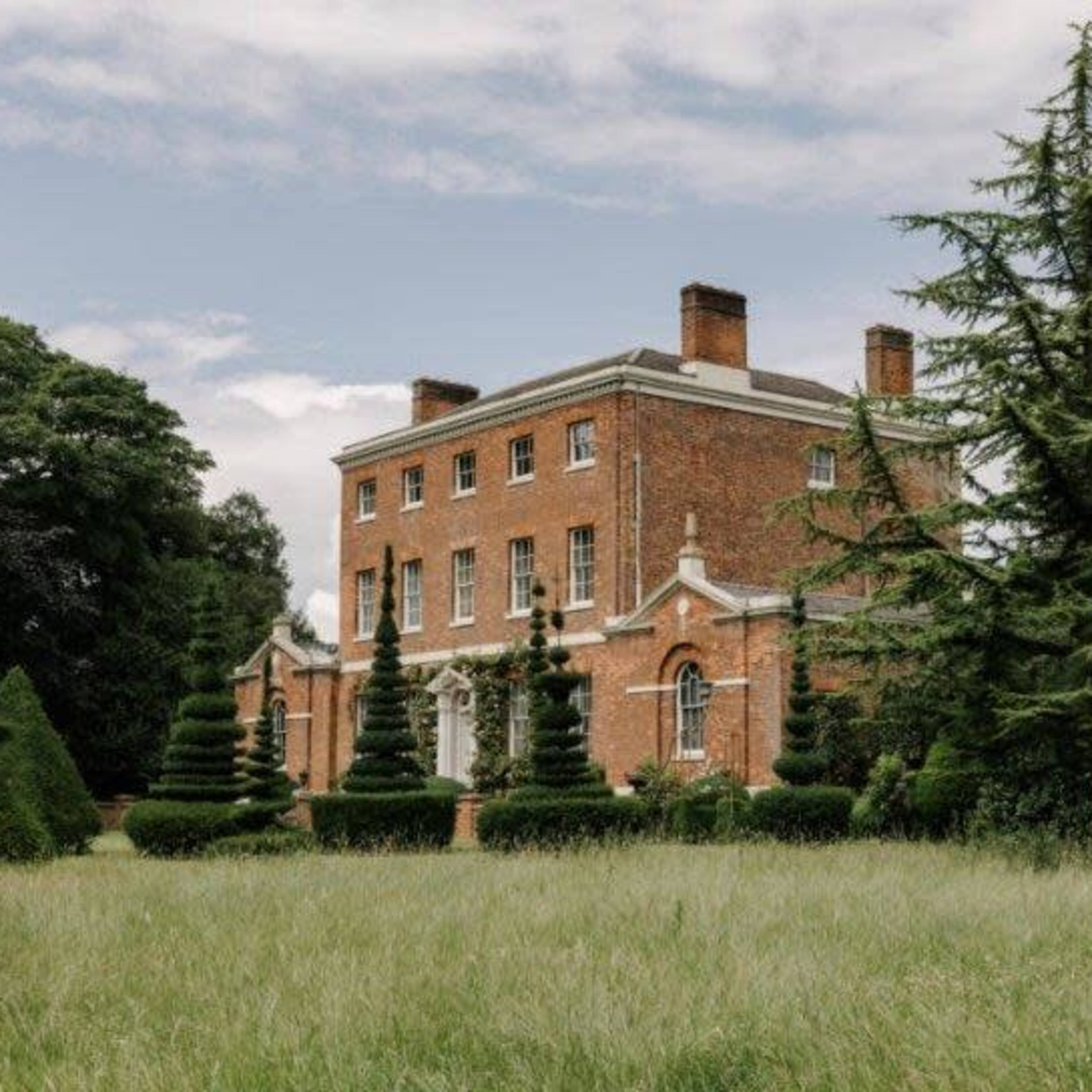 A large brick mansion with symmetrical windows is surrounded by manicured lawns and conical topiary trees.
