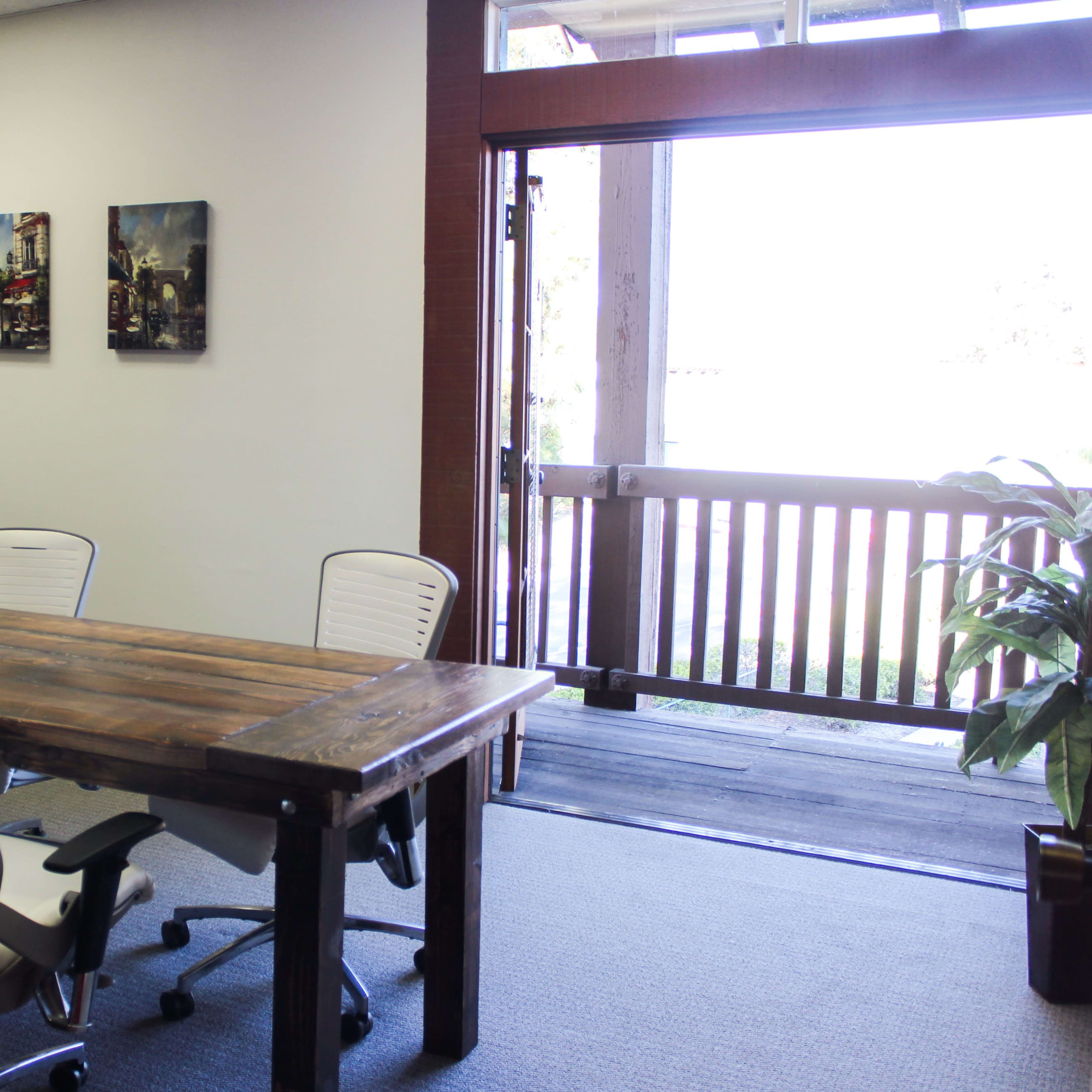 A conference room features a wooden table surrounded by white chairs, with a door leading to an outdoor area visible in the background.