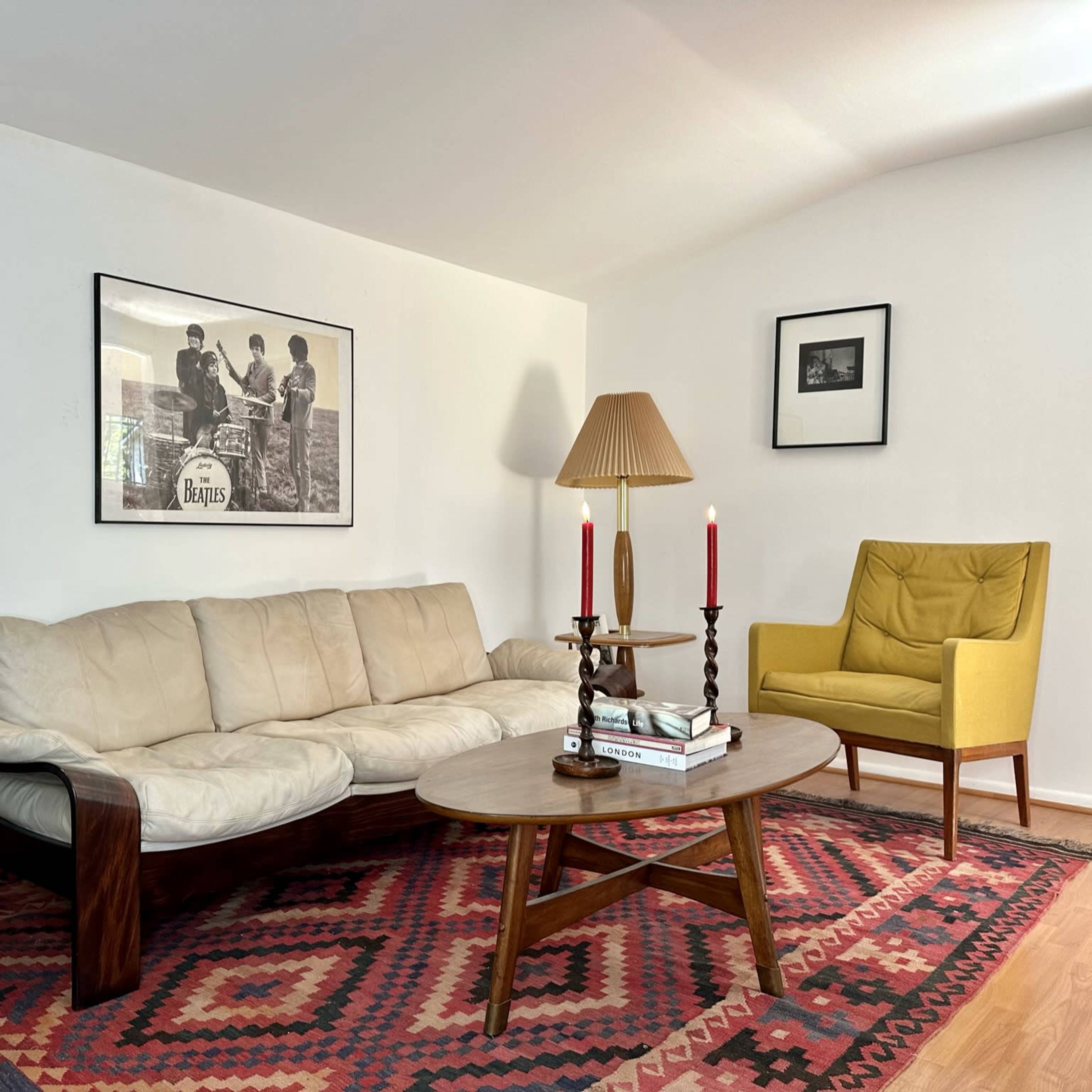 A living room with a beige couch, a wooden coffee table, a yellow armchair, and a decorative rug, accompanied by framed pictures on the walls.