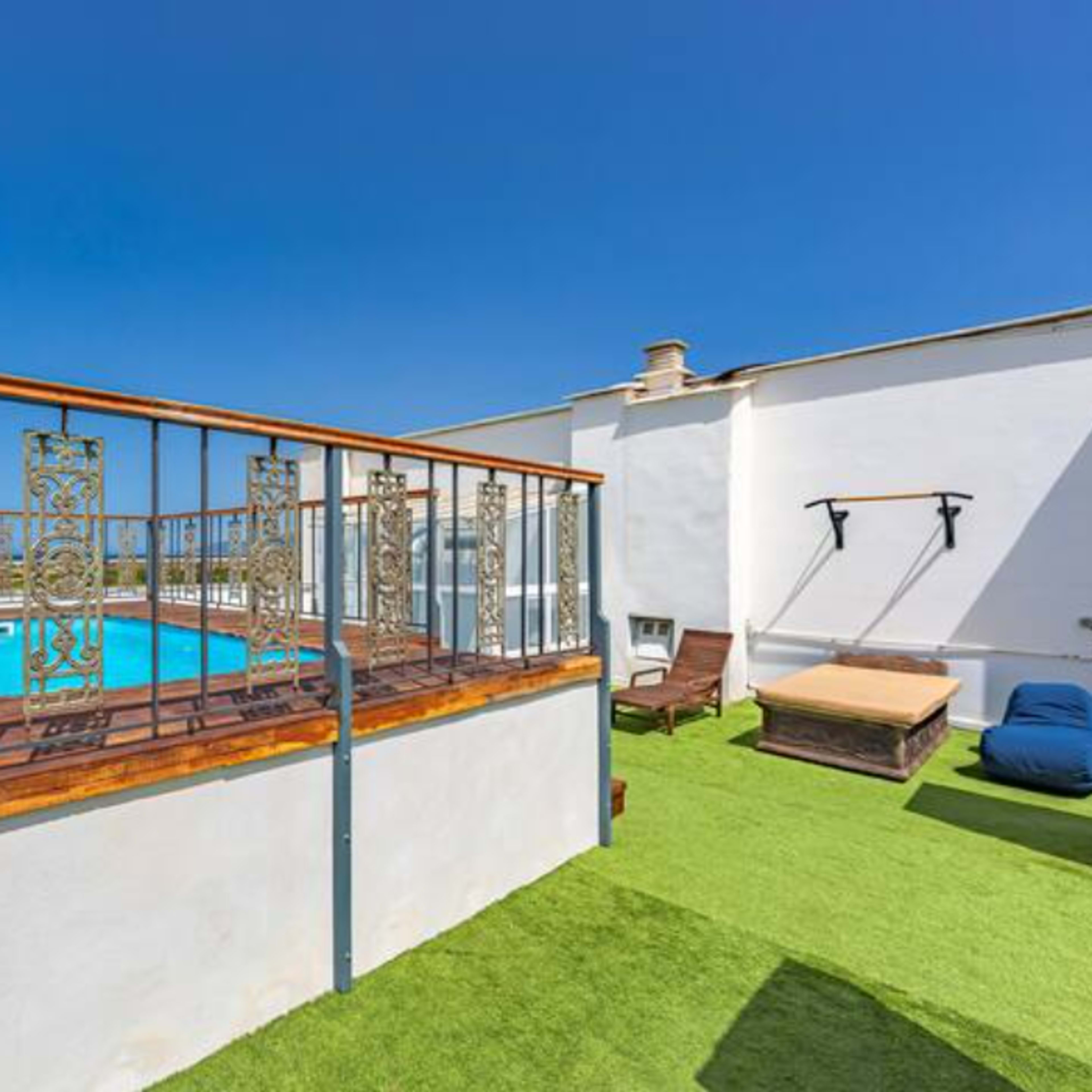 A rooftop area featuring a small pool surrounded by a wooden deck and metal railing, with green artificial grass and lounge chairs.