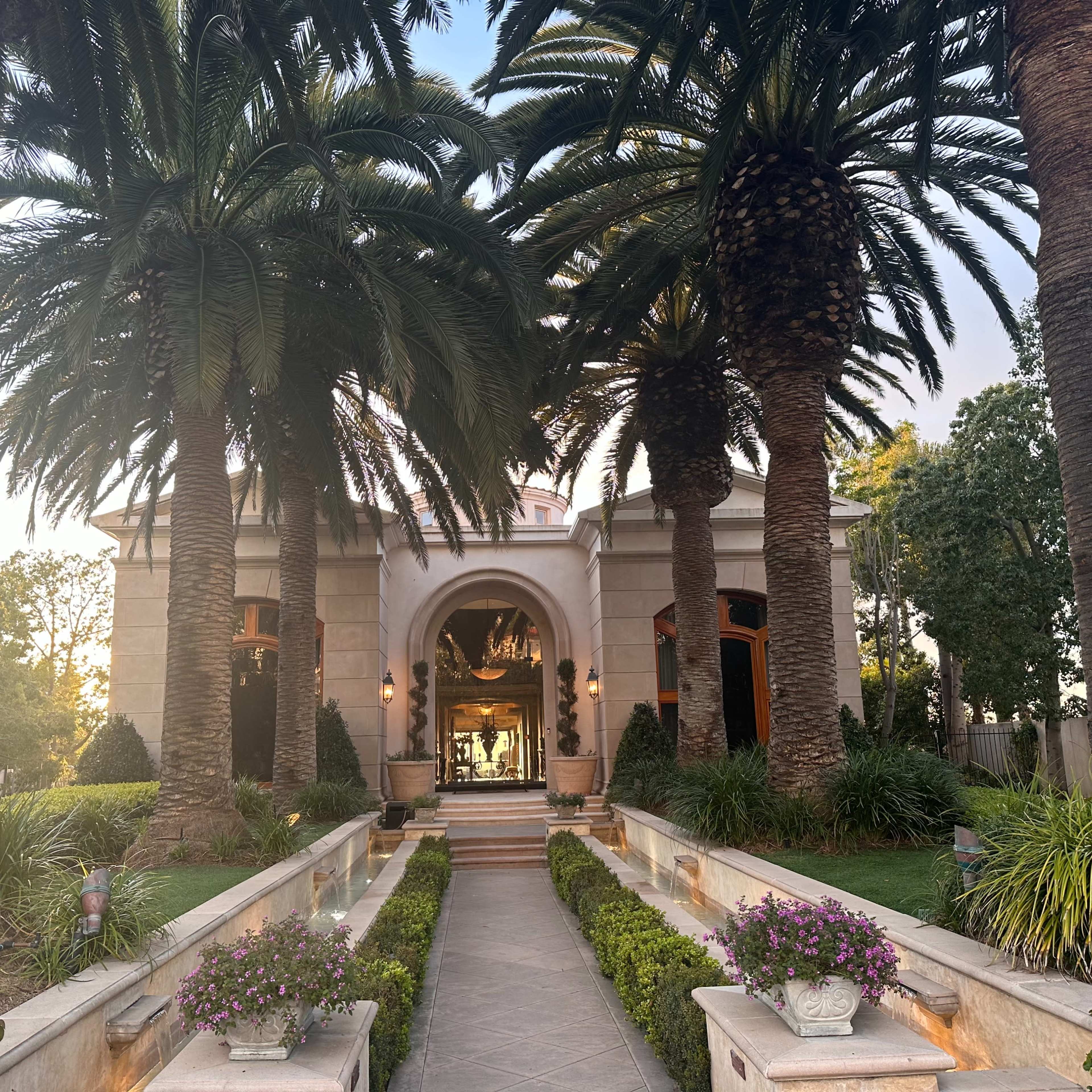 A grand house with a large entrance is framed by tall palm trees and has a landscaped pathway lined with flowers.