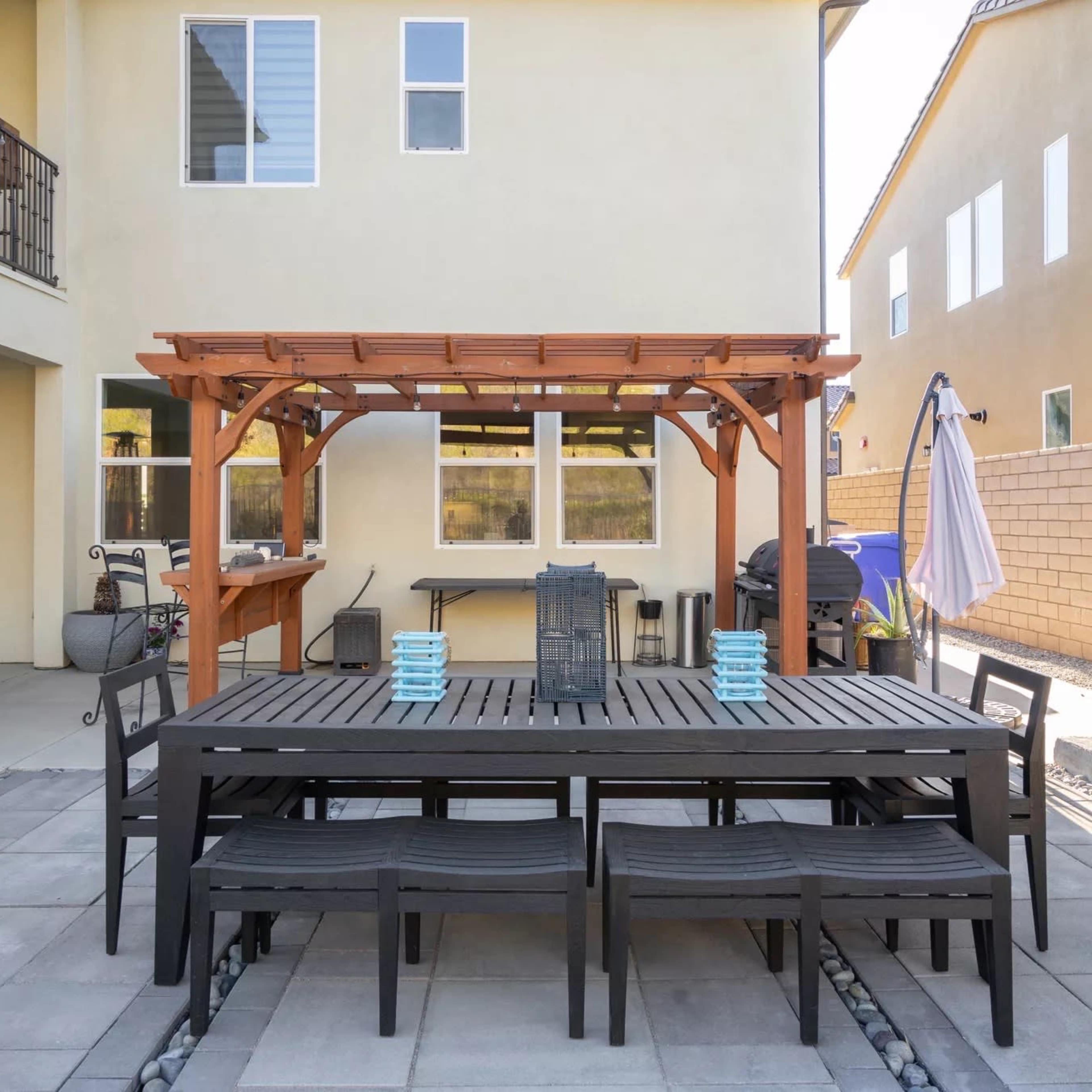 A patio area with a wooden pergola over a large dining table and chairs, adjacent to a modern home with multiple windows.