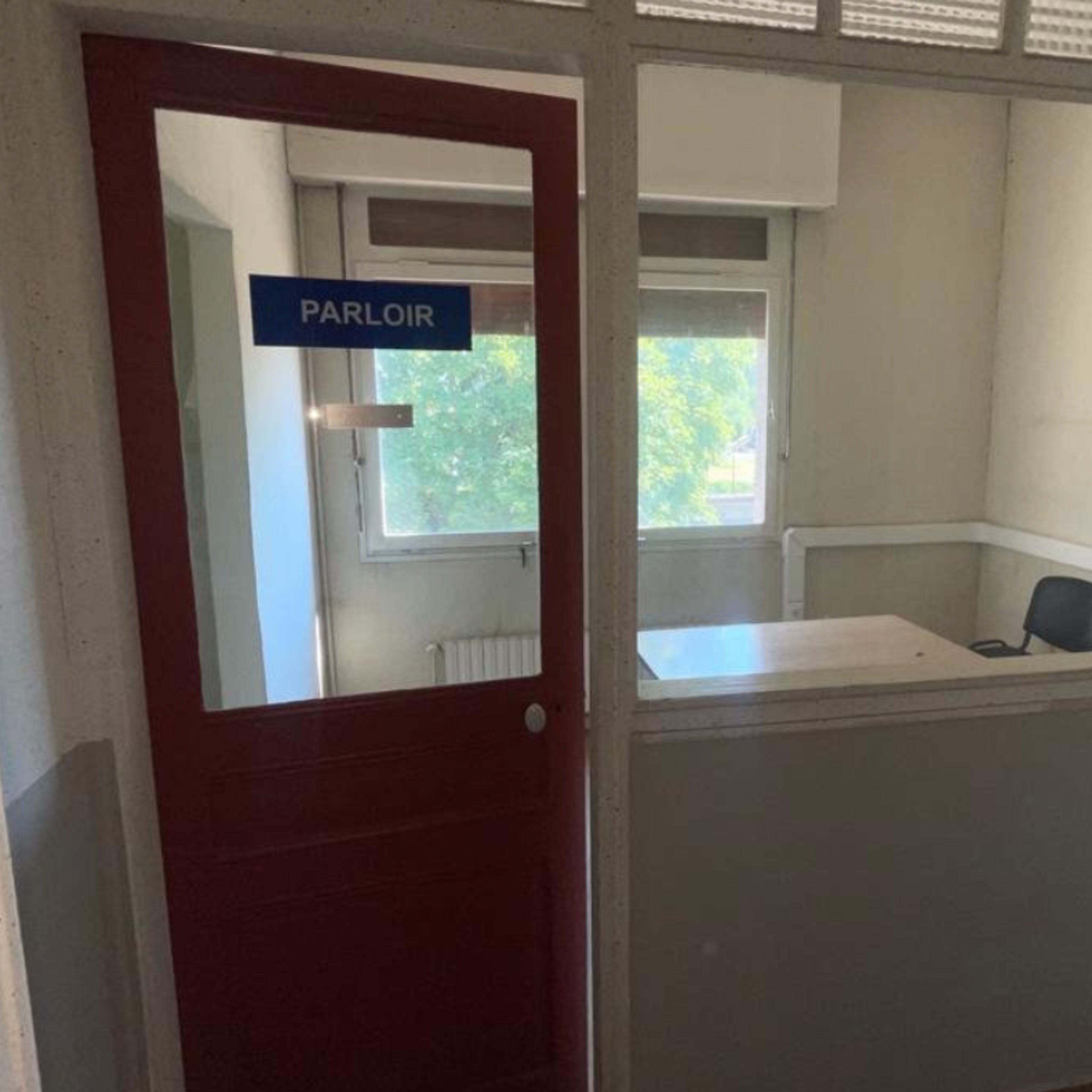 A small office with a glass wall and a red door labeled "PARLOIR," featuring a table and a single chair inside.