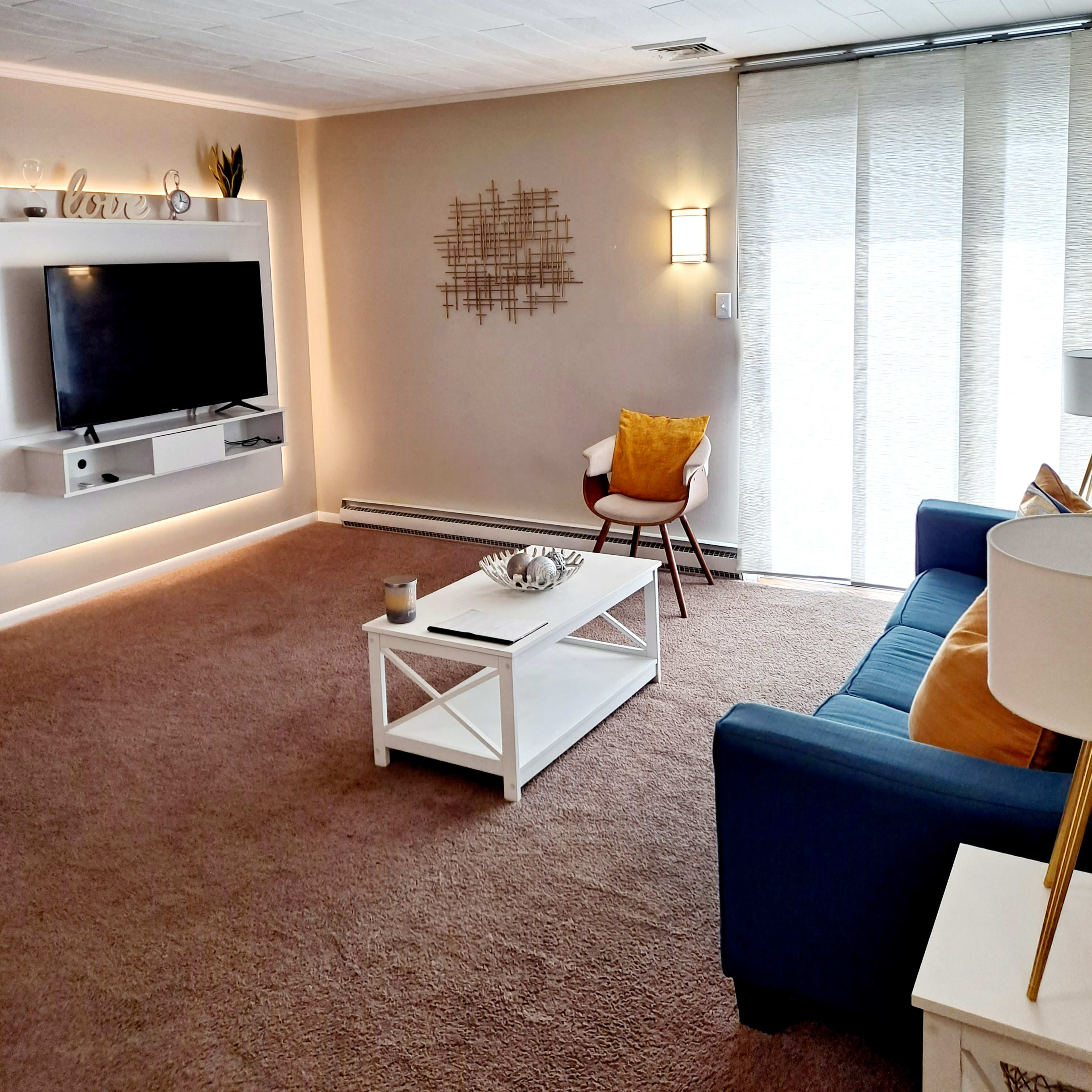 A cozy living room with a blue sofa, a white coffee table, a television mounted on the wall, and large windows covered with light, sheer curtains.