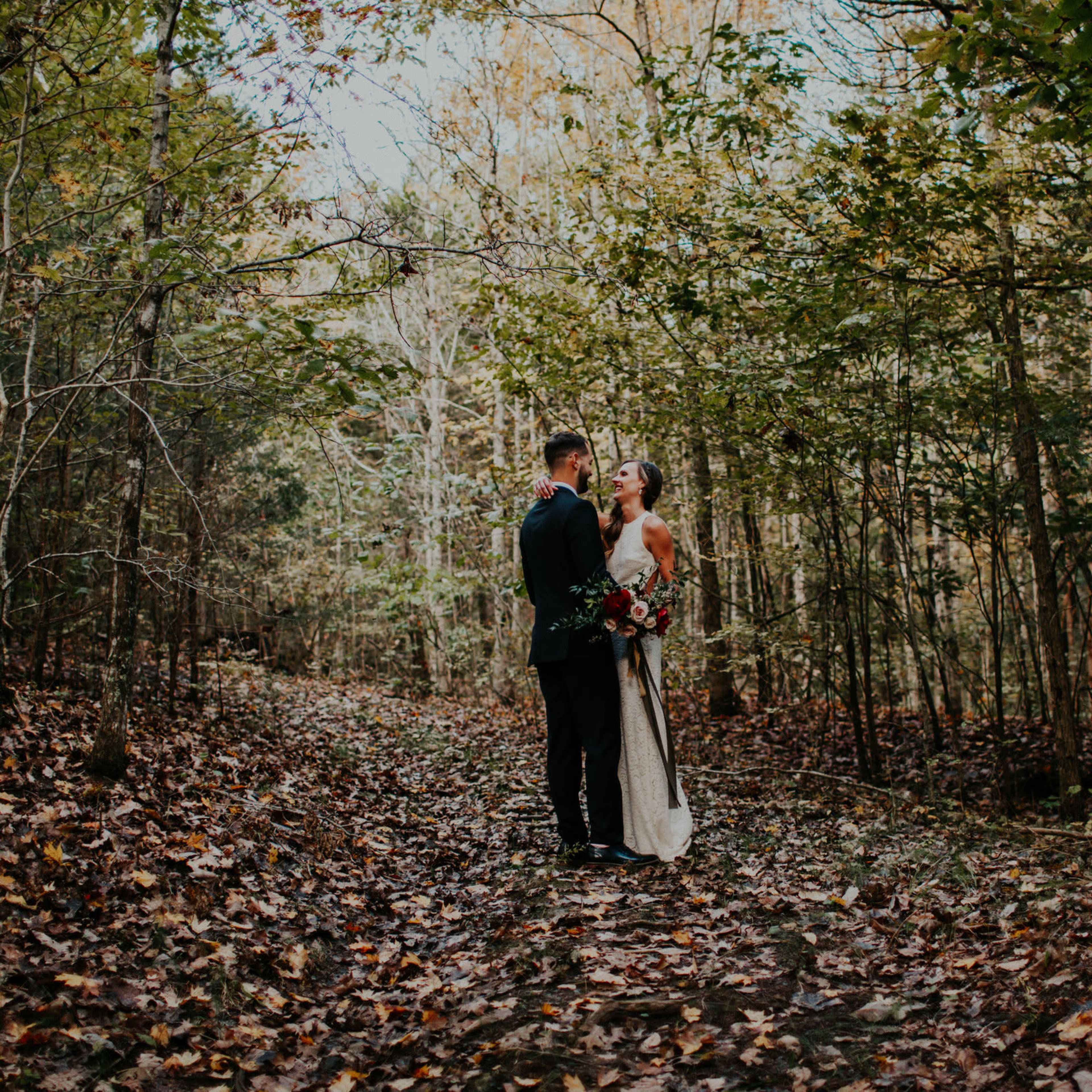 A couple stands together on a leaf-covered path in a forest, surrounded by trees.