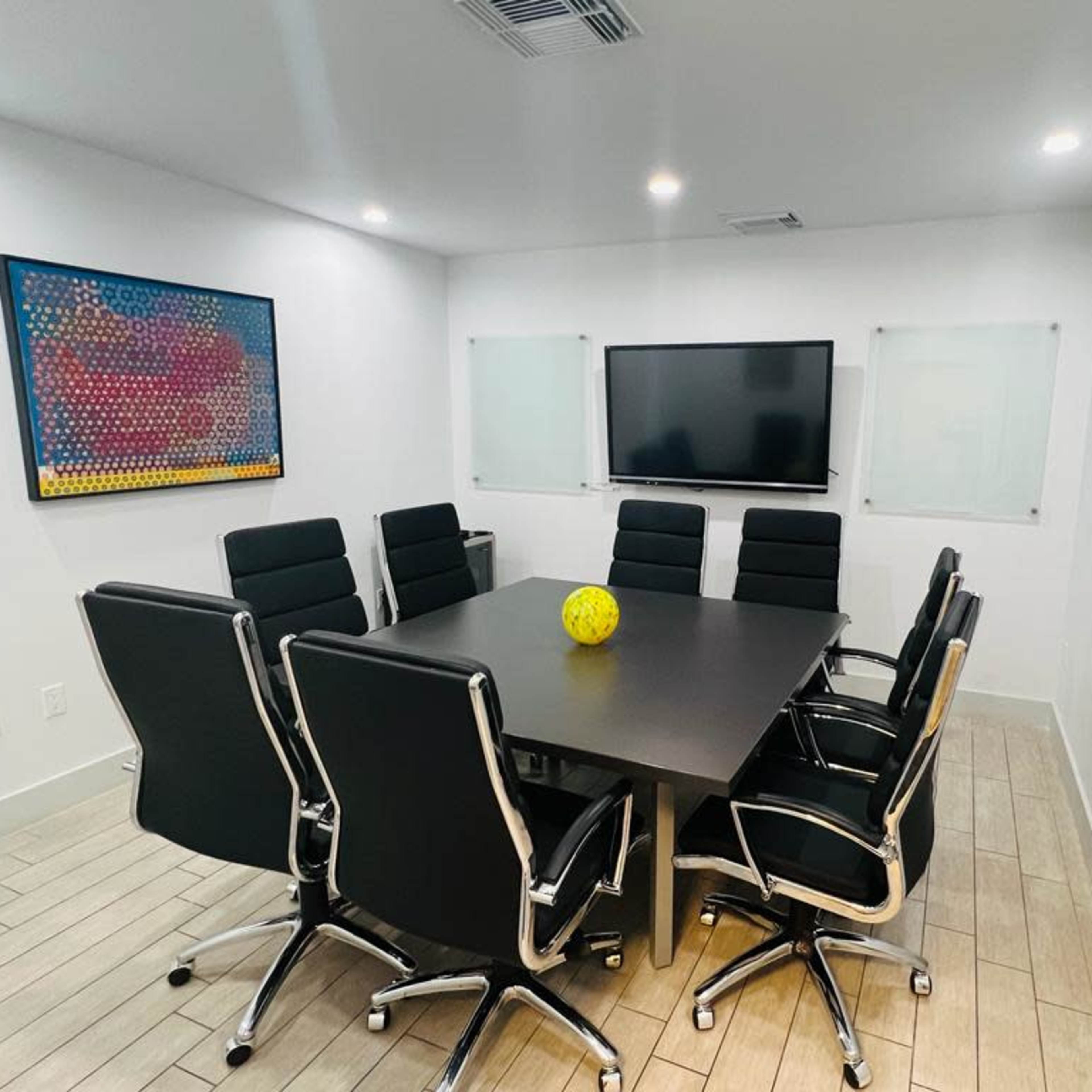 A modern conference room features a large black table surrounded by eight black chairs and a yellow decorative item at its center, with a television and whiteboards on the walls.