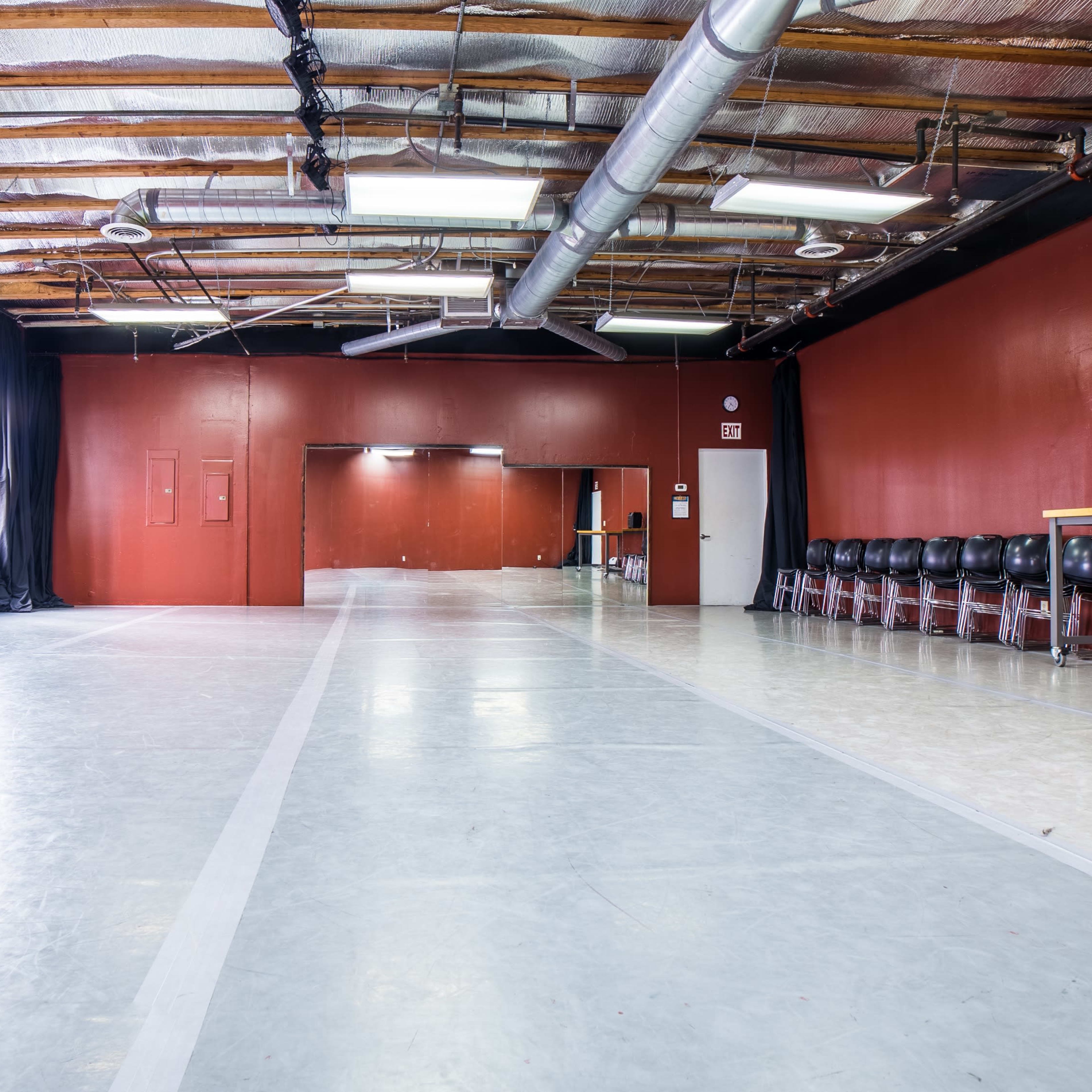 An empty studio space features a polished floor and red walls, with a row of black chairs along one side.