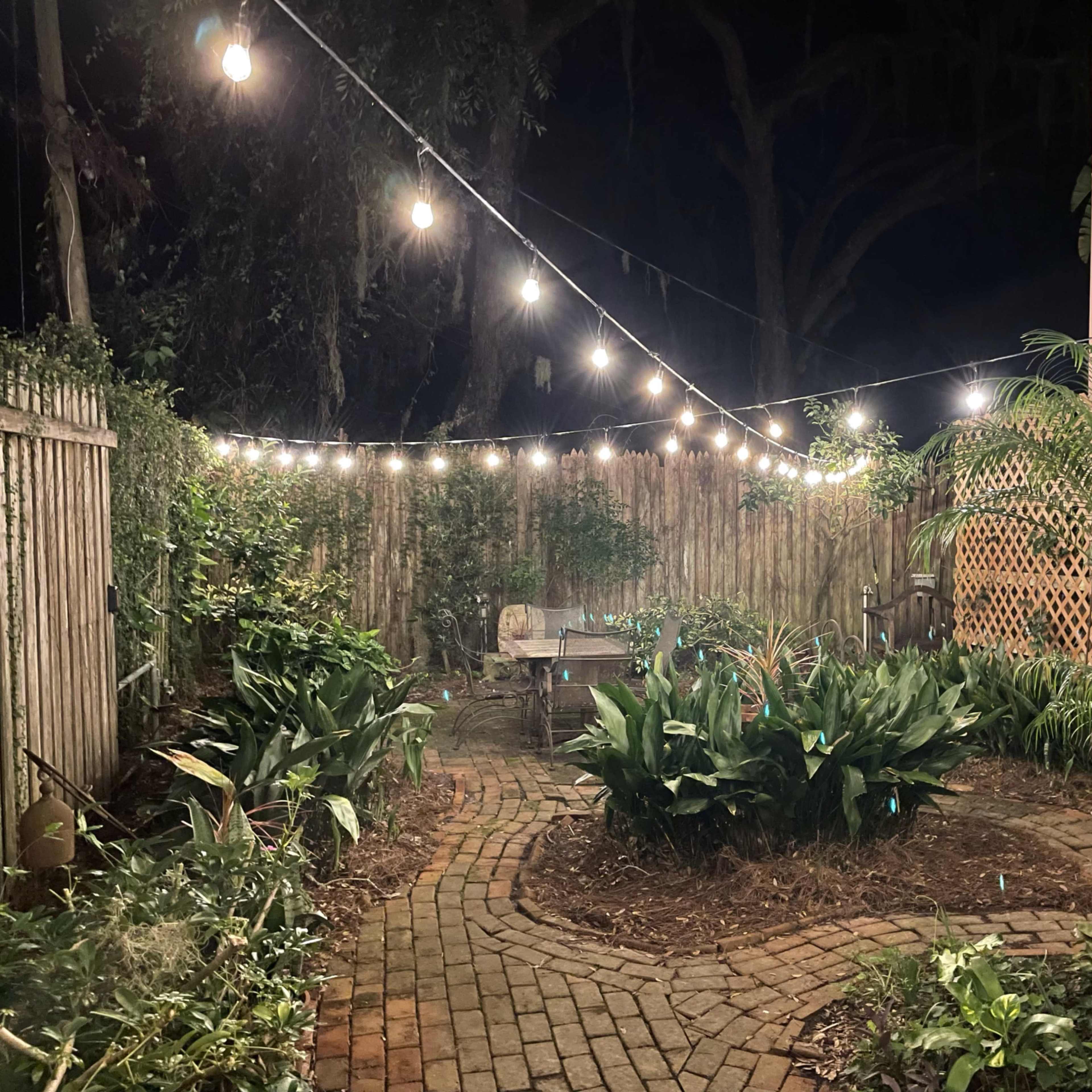 The scene shows a brick path winding through a garden illuminated by string lights at night.