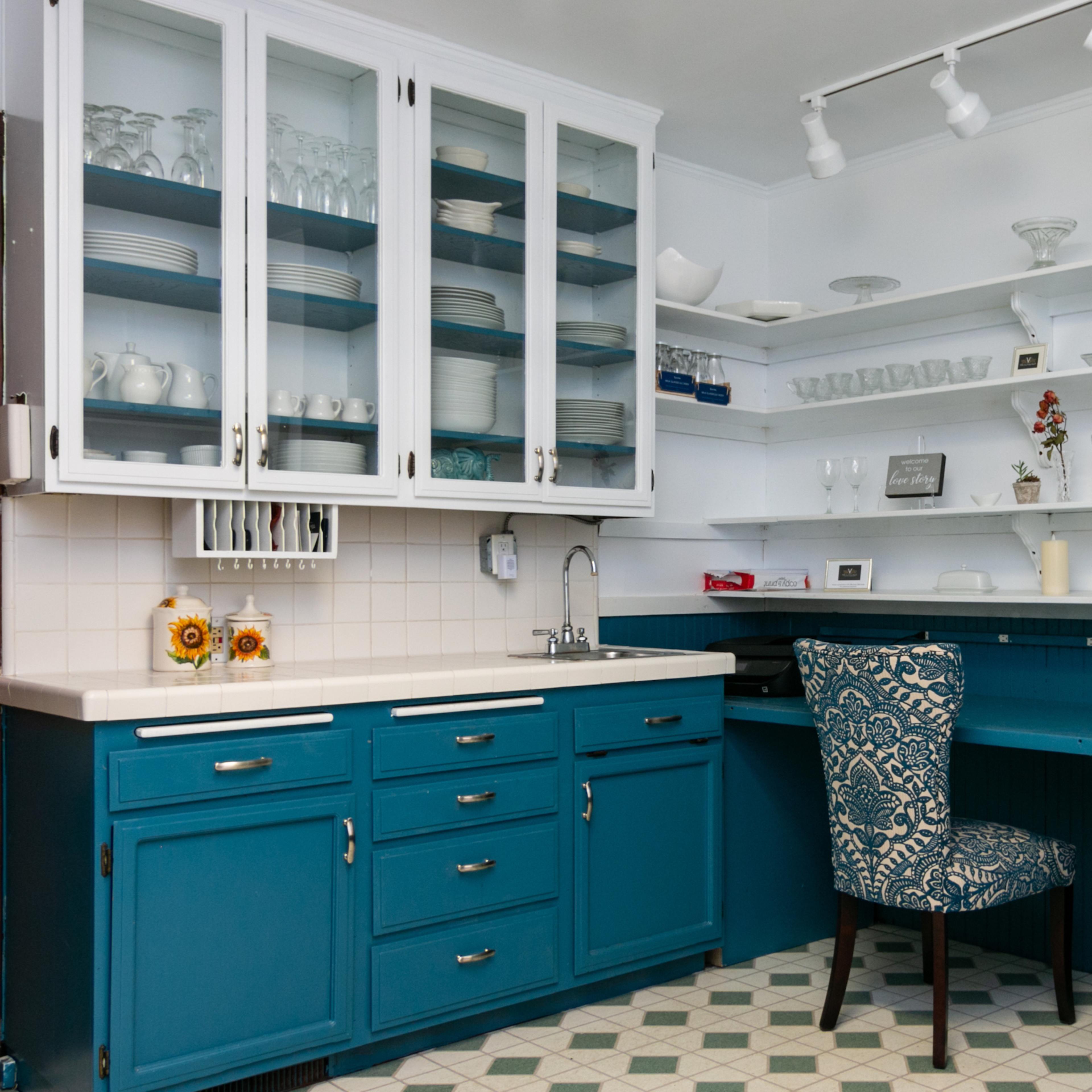 A modern kitchen with blue cabinetry, a desk area, and glass-front upper cabinets displaying dishware.