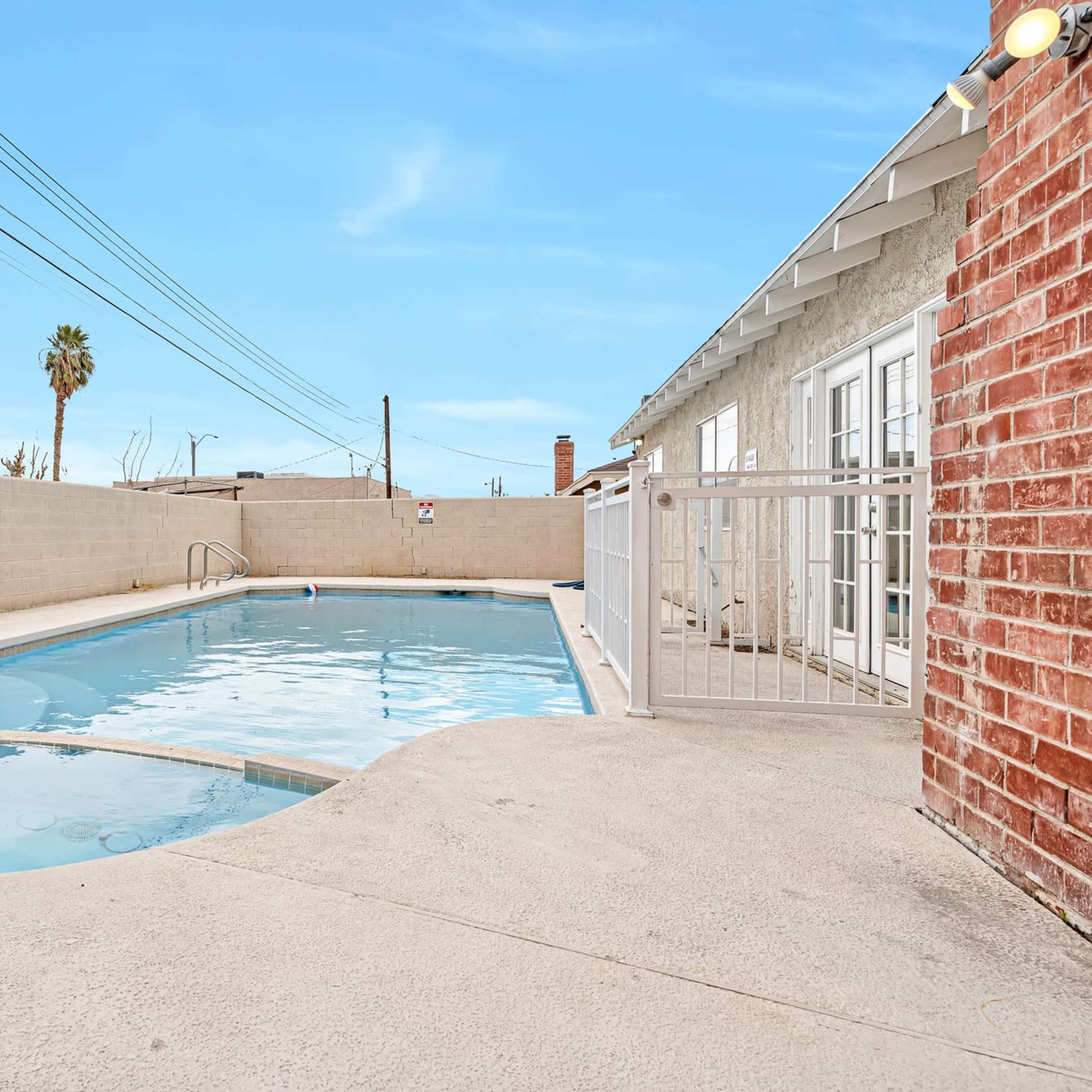 A swimming pool surrounded by concrete, with a fence, palm trees in the background, and a house on one side.